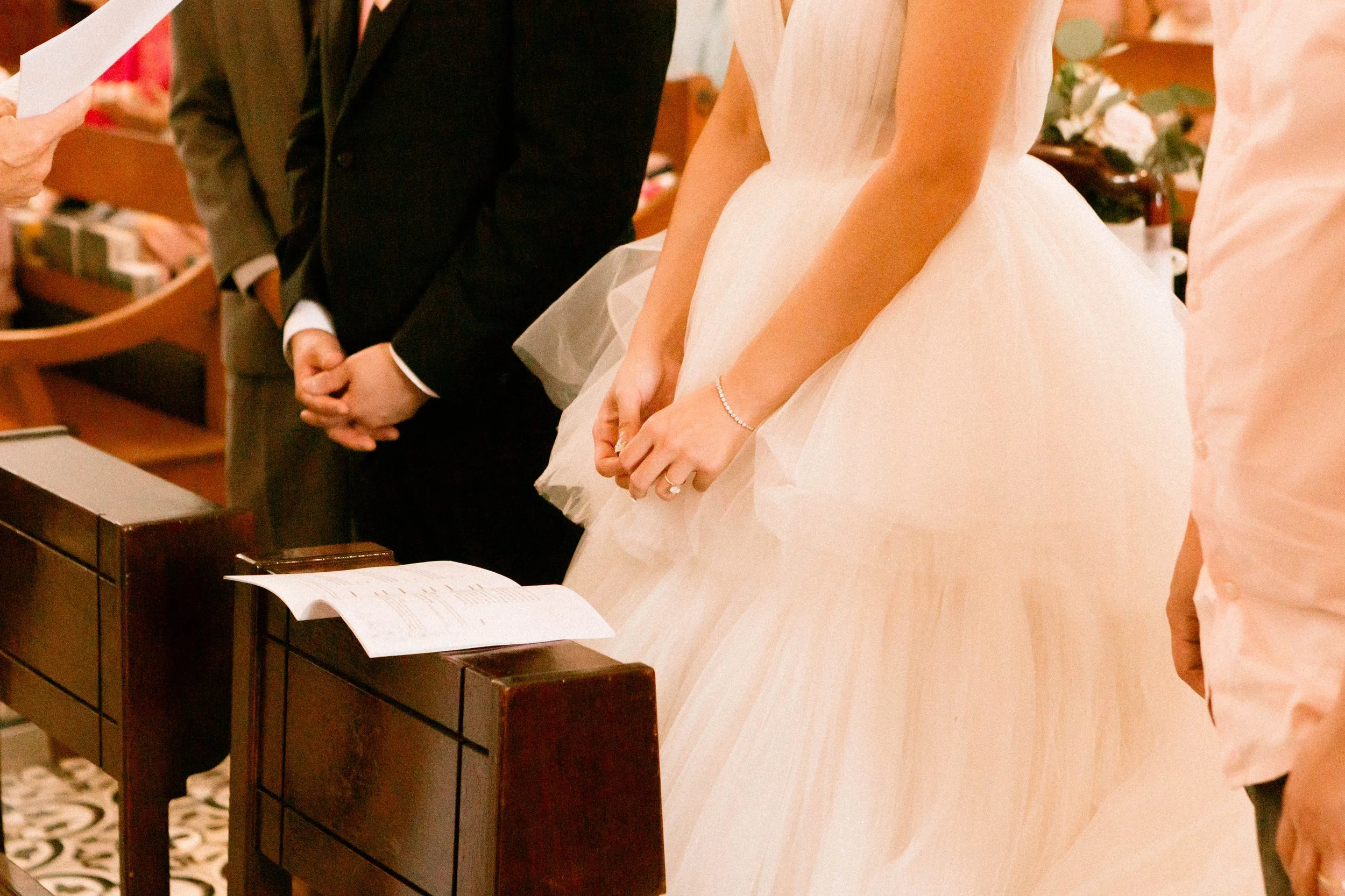 Close-up of a bride in a white wedding gown and a groom in a black tuxedo standing during a wedding ceremony, with a person on the left holding a document.