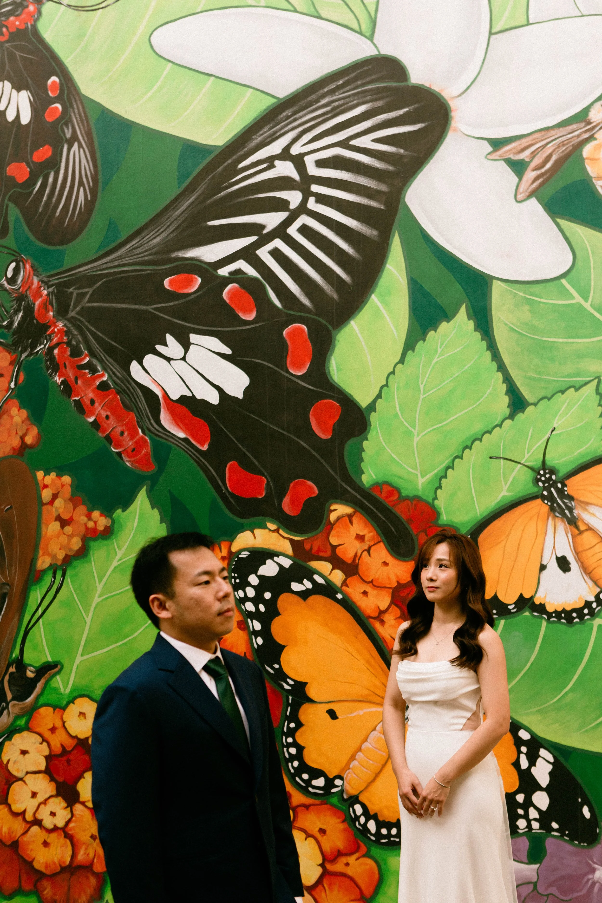 A woman in a white dress and a man in a dark suit stand in front of a colorful mural of butterflies and flowers.