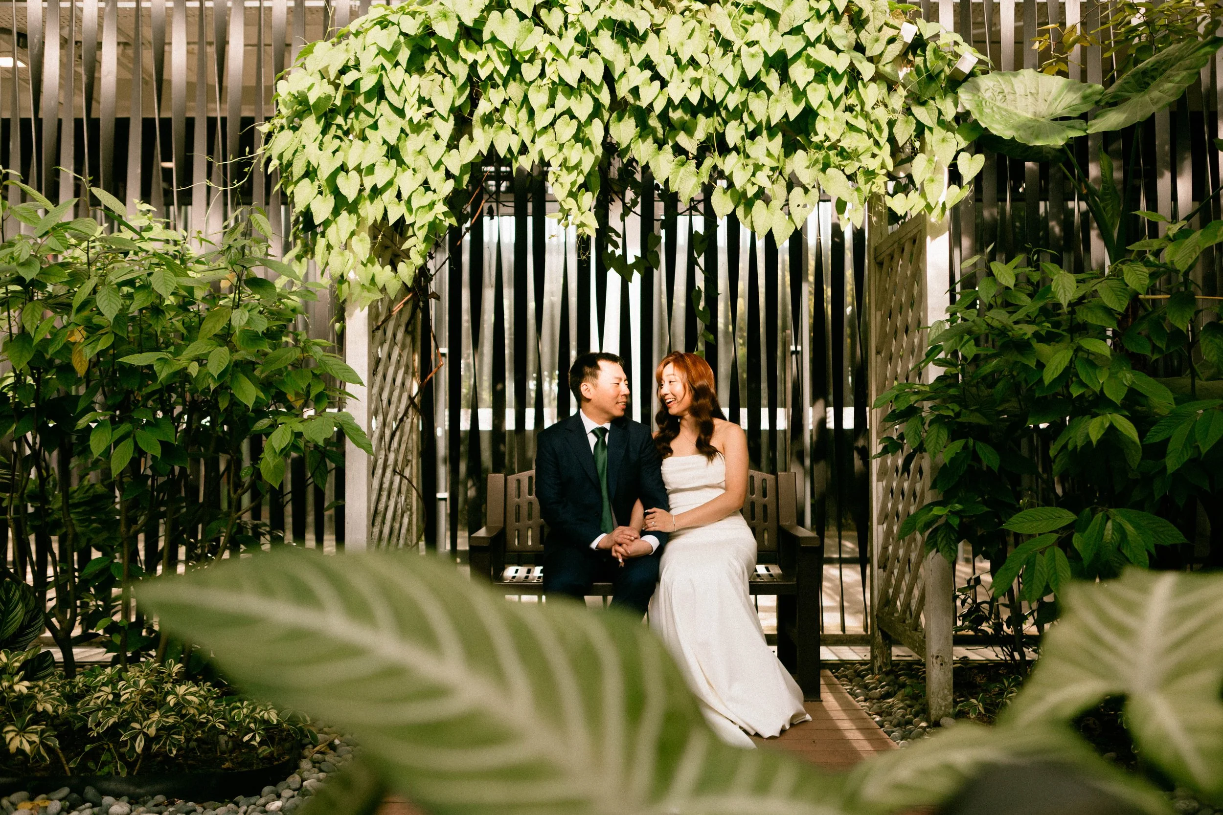 A couple in formal attire sitting on a wooden bench inside a lush, green garden with hanging and potted plants, smiling and looking at each other.