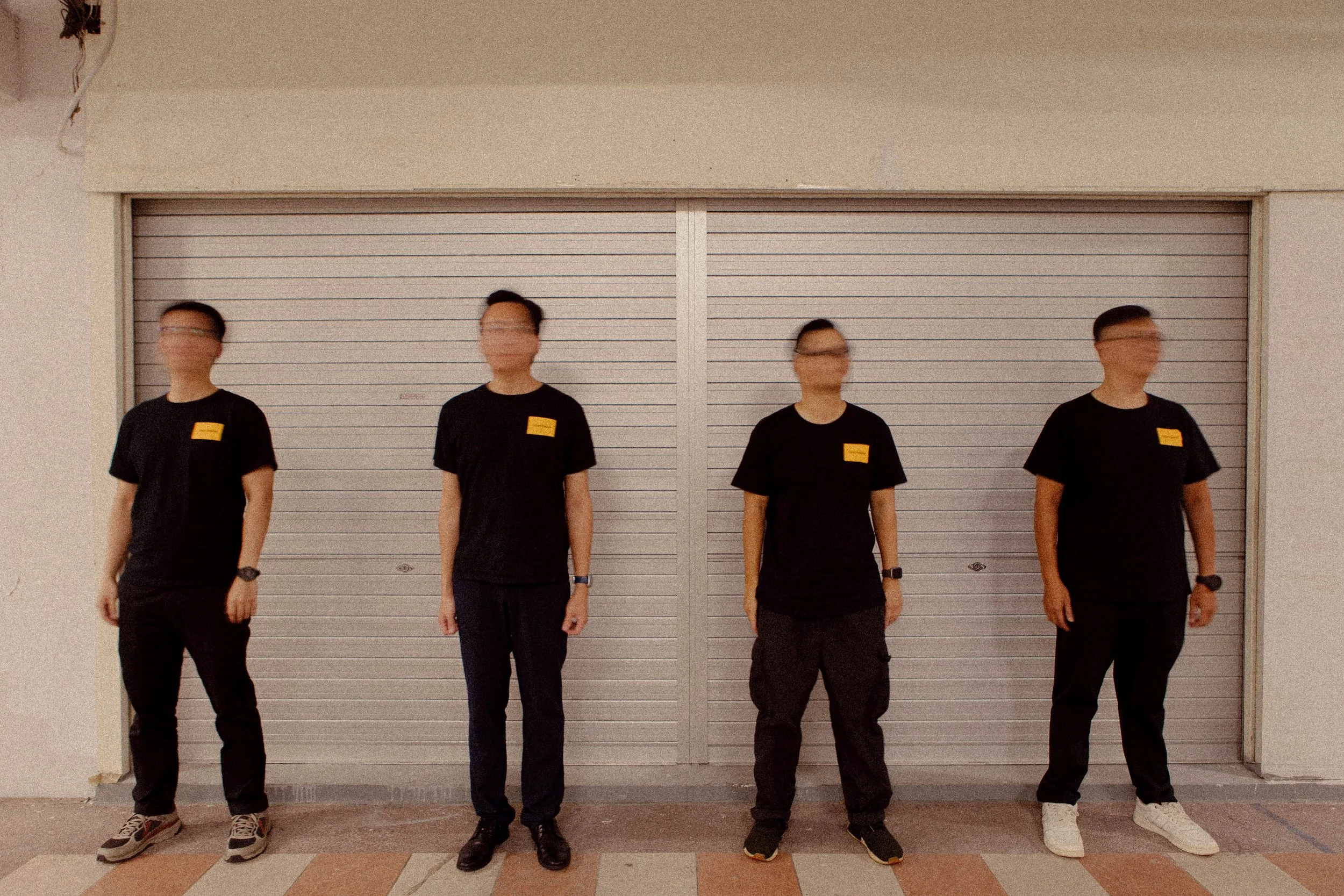 Four men wearing black t-shirts with yellow square patches standing in front of a closed gray metal store shutter.