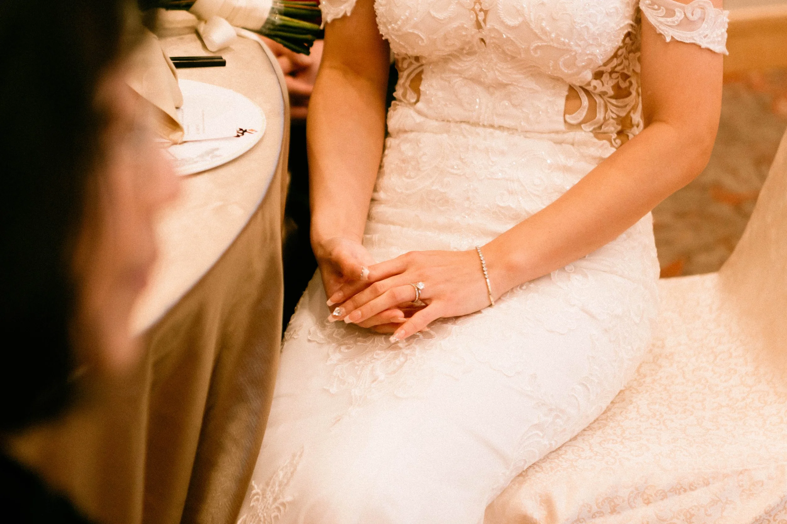 Close-up of a bride sitting with her hands resting on her lap, wearing a white lace wedding dress, with her hands clasped, showing her jewelry including a ring and bracelet, at a wedding reception.