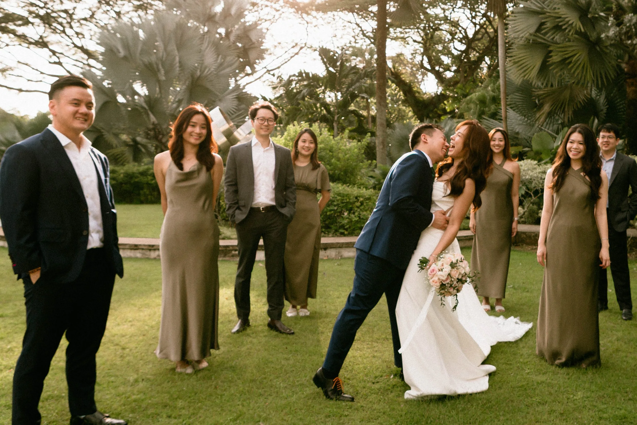 A group of people at a wedding, with a bride and groom in the center, surrounded by friends in formal attire, outdoors on a grassy area with trees.