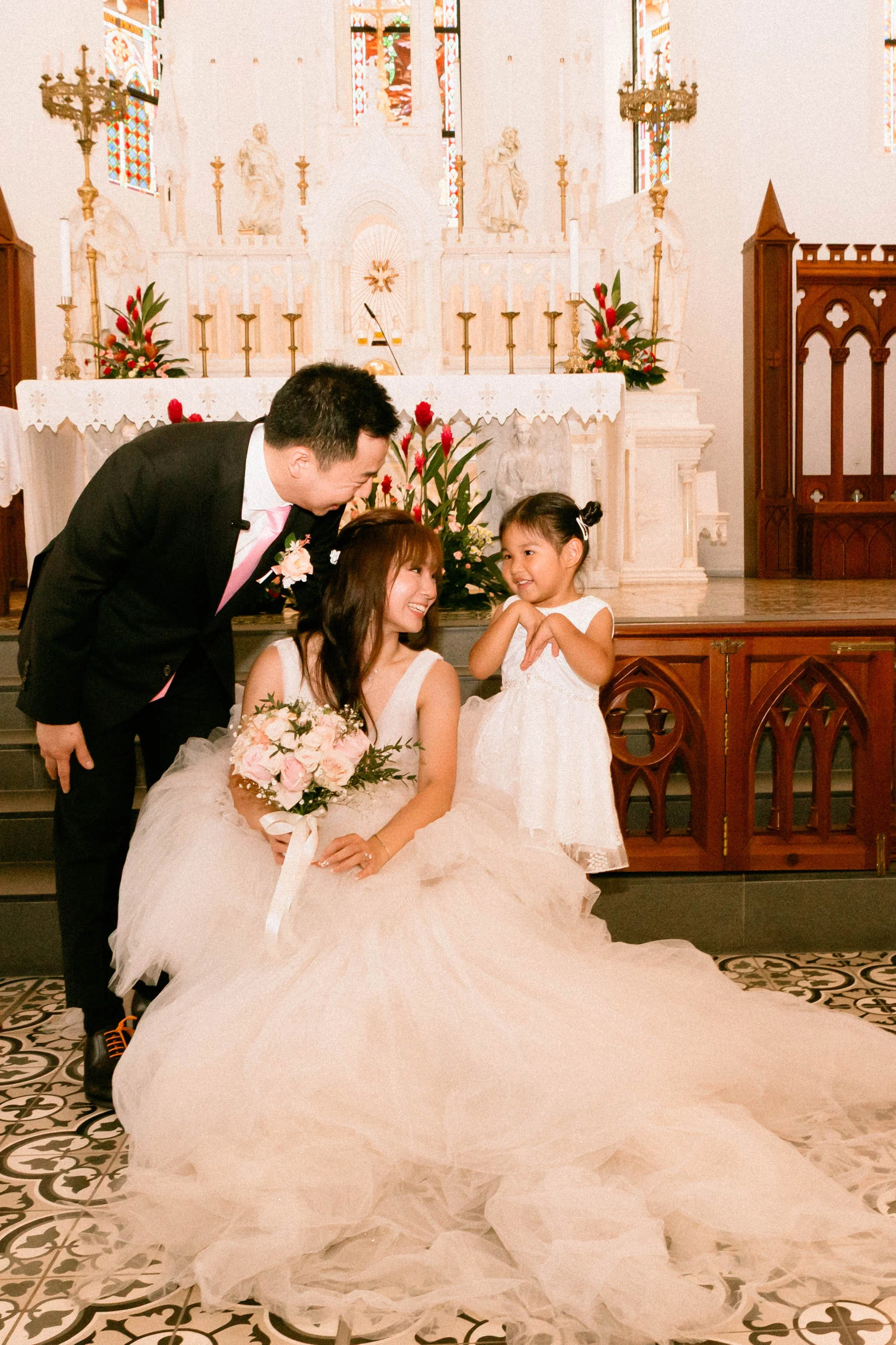 A bride, groom, and young girl pose inside a church with stained glass windows and altar decorations. The bride is seated in a white wedding gown, holding a bouquet of pink and white flowers. The groom is leaning toward the bride, and the young girl