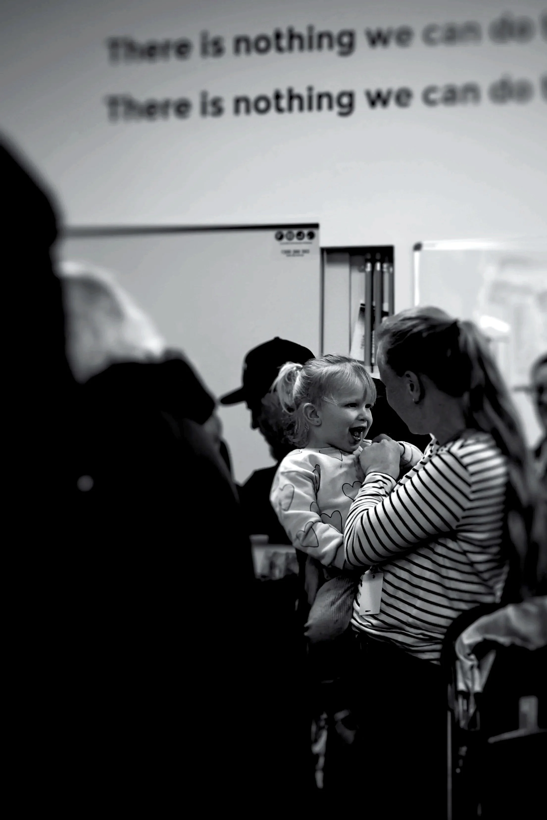 A young girl laughing and smiling while interacting with an adult in a striped shirt in a room with several people, some wearing hats, and a whiteboard or screen in the background.