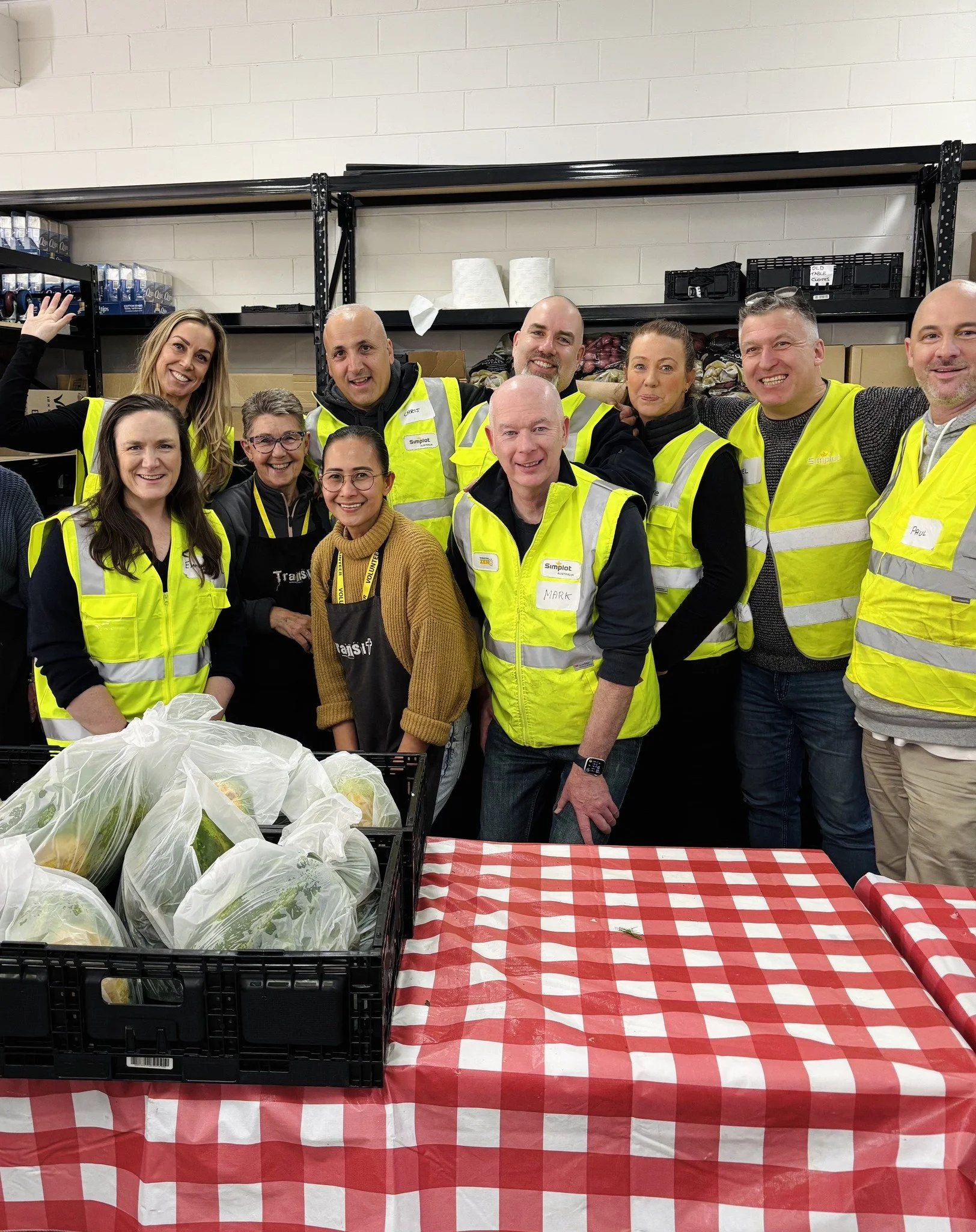 Group of people wearing yellow safety vests smiling and posing together in a warehouse, standing behind a table with a red and white checkered tablecloth and bags of produce.