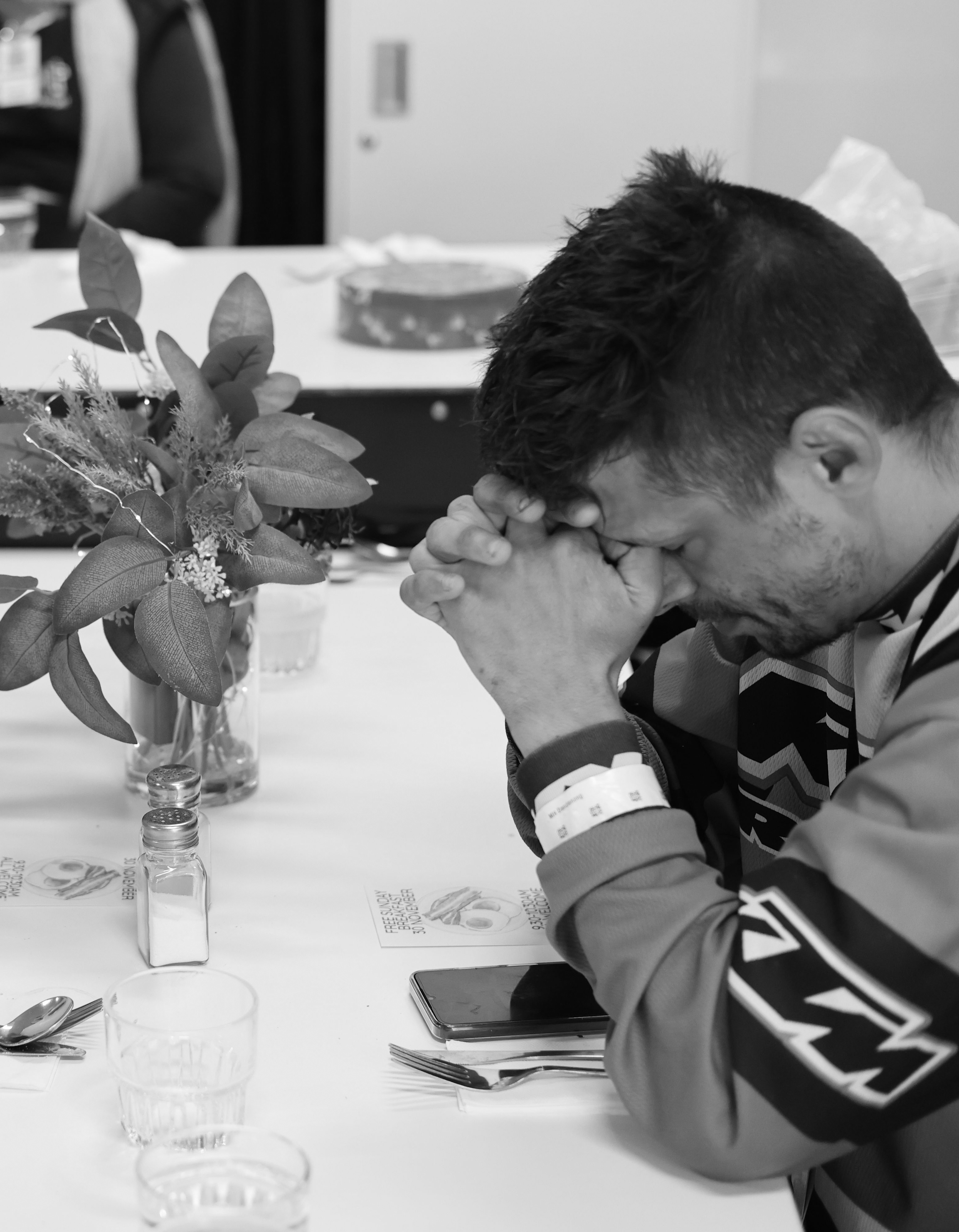 A man with short dark hair and a beard praying with his hands clasped and head bowed at a table in a dining area, with flowers, salt shakers, utensils, a smartphone, and a glass of water in front of him.