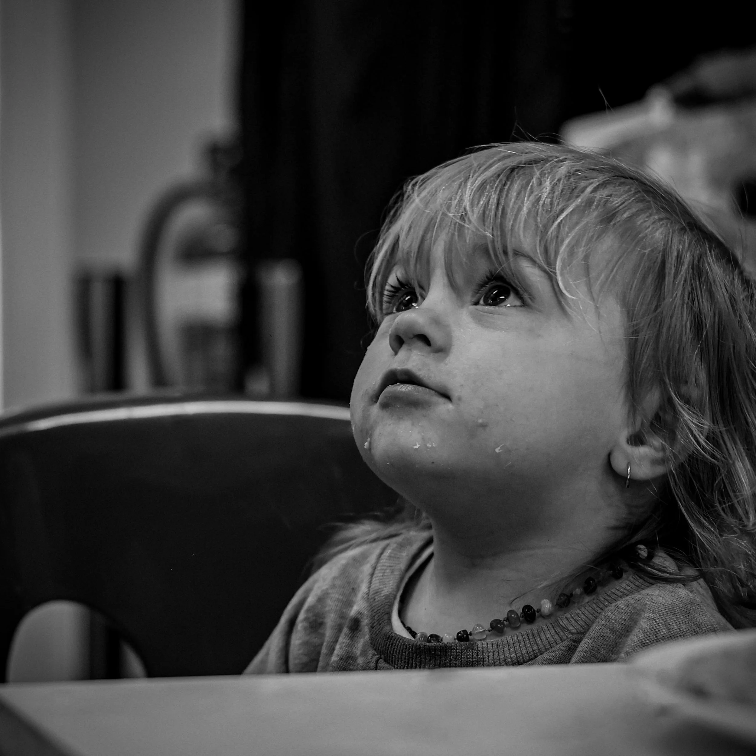 A young girl with light-colored hair, wearing a necklace with beads, looking upward with a curious expression in a grayscale photograph.
