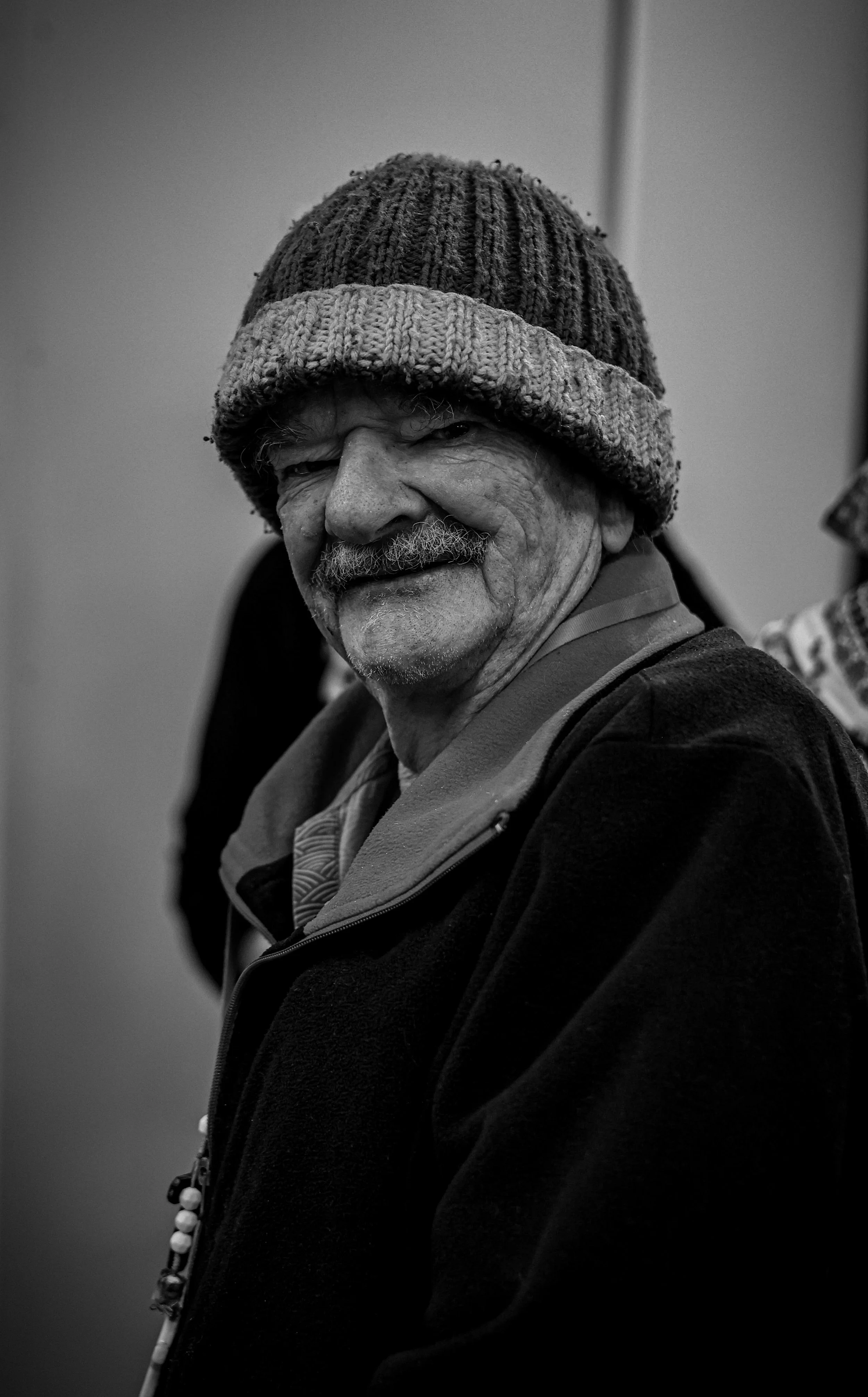 A black and white photo of an elderly man wearing a knitted beanie, smiling with a mustache and a goatee, dressed in a jacket.