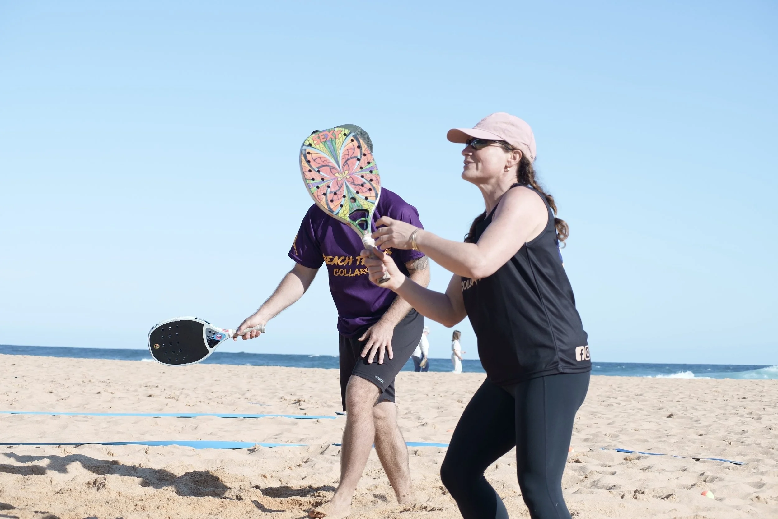 People playing Beach Tennis on a sandy beach with a rainbow in the sky and the ocean in the background.