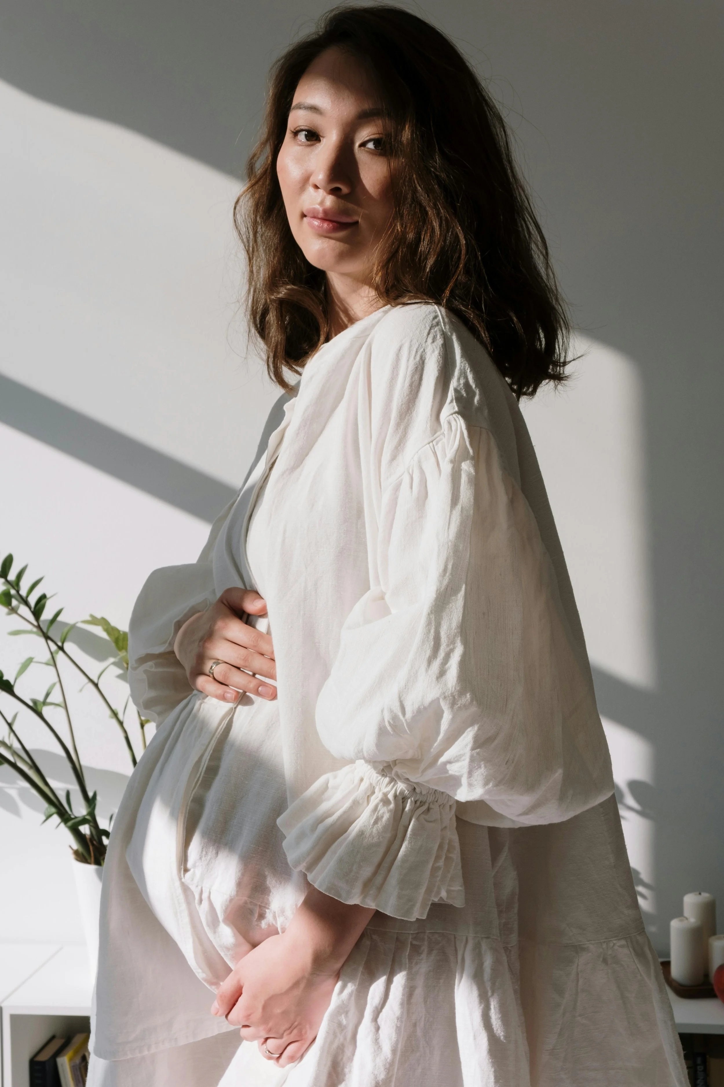 A woman with long, wavy brown hair wearing a loose white blouse, standing in front of a white wall with shadows cast on it. She has a gentle smile and is holding her hands together at her waist. Some plants are visible in the foreground, and candles are seen in the background.