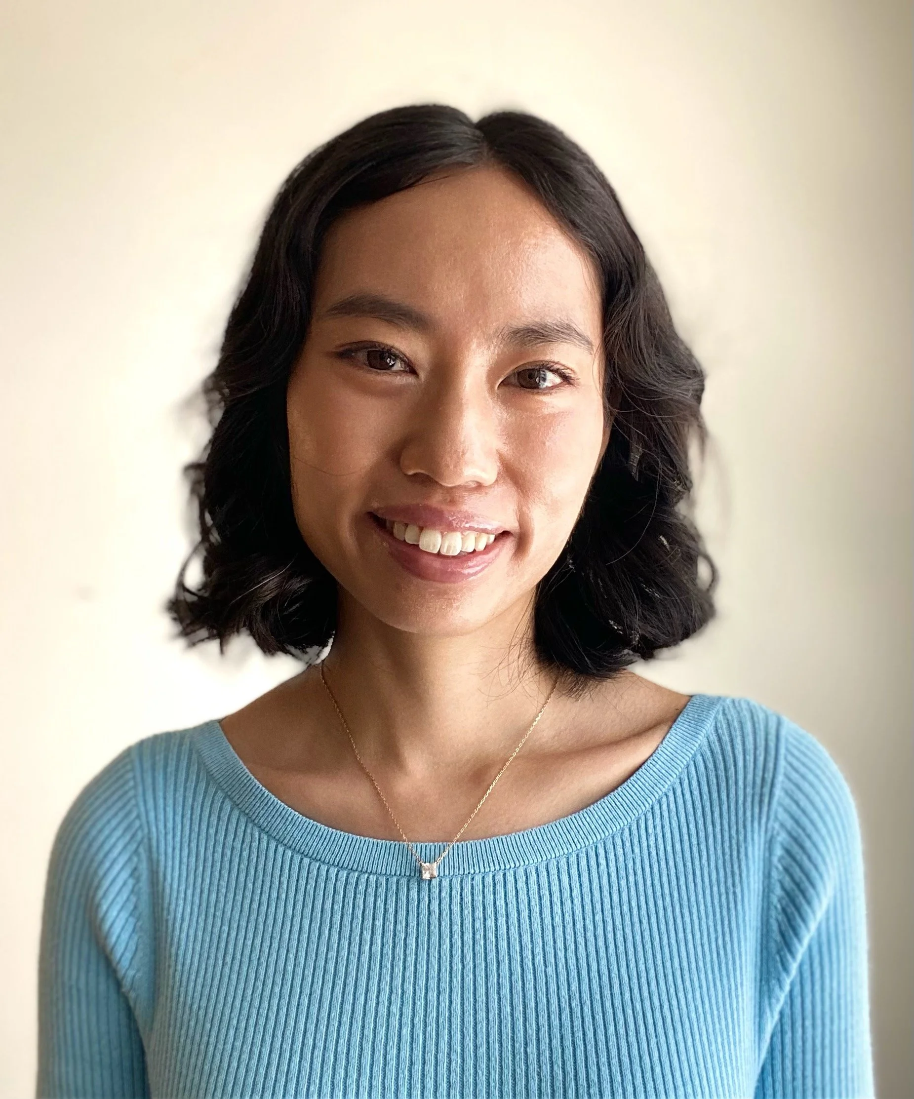 Close-up of a smiling woman with dark, wavy hair wearing a blue ribbed sweater and a delicate necklace with a heart-shaped pendant, standing against a plain beige background.
