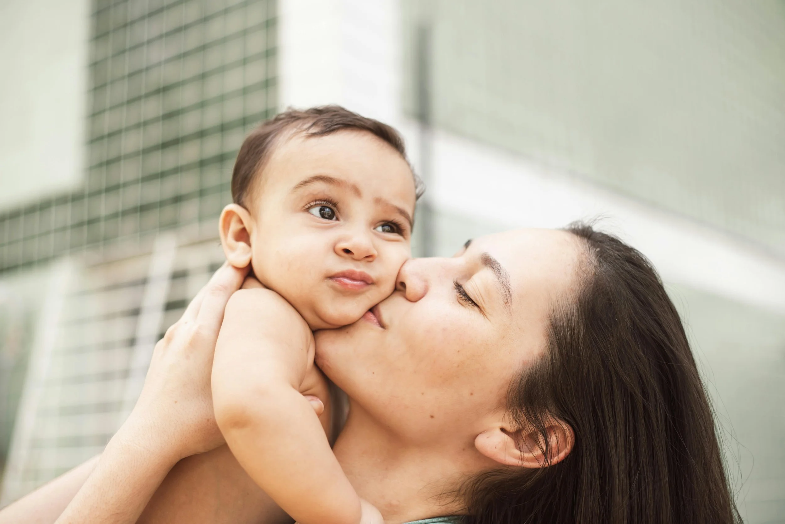 A child being kissed on the cheek by an adult woman, possibly their mother, outdoors near a building with a grid-like fence.
