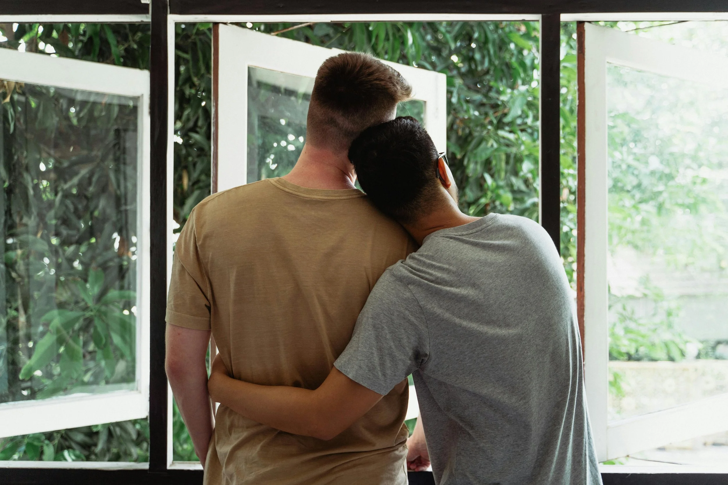 Two people hugging near an open window with a view of green foliage outside.