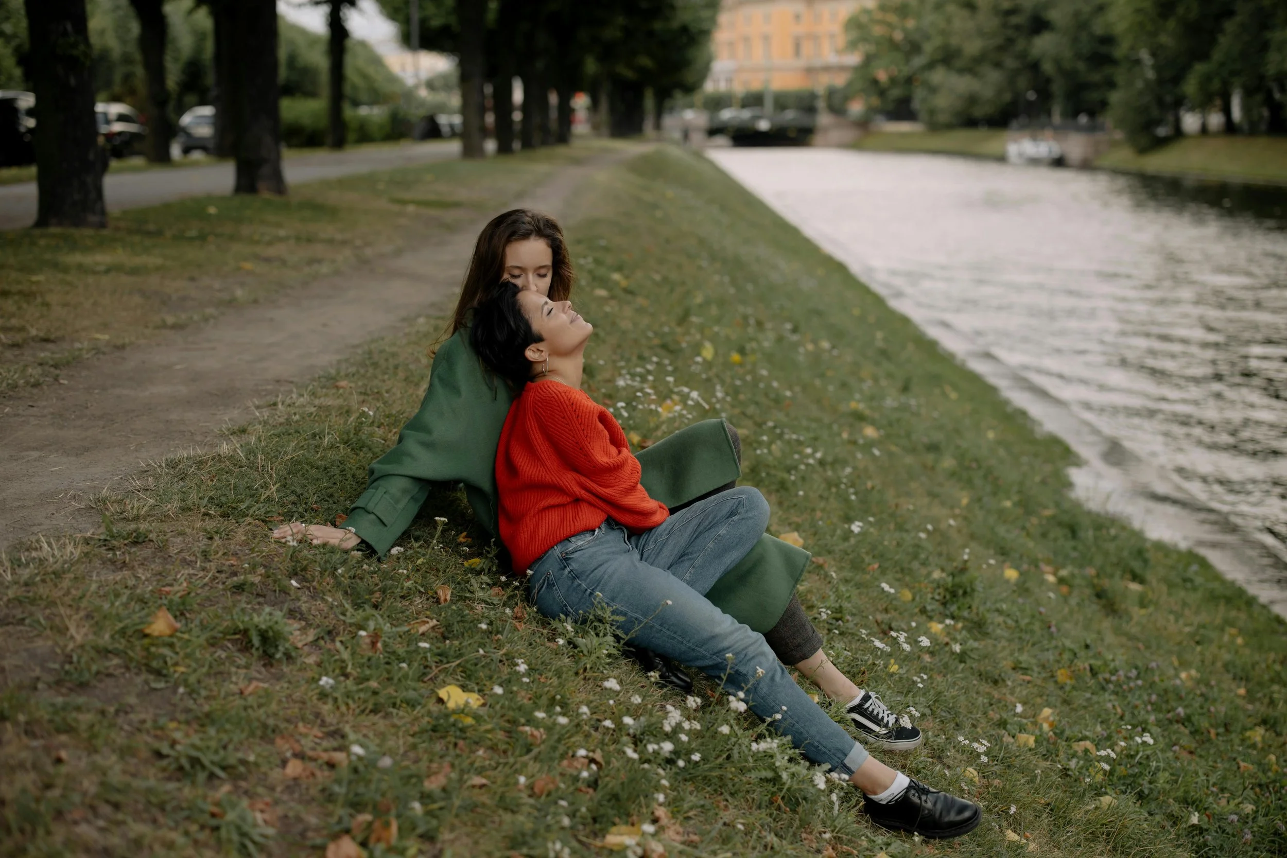 Two women sitting on a grassy riverbank, embracing and smiling during autumn in a park with trees and a city building in the background.
