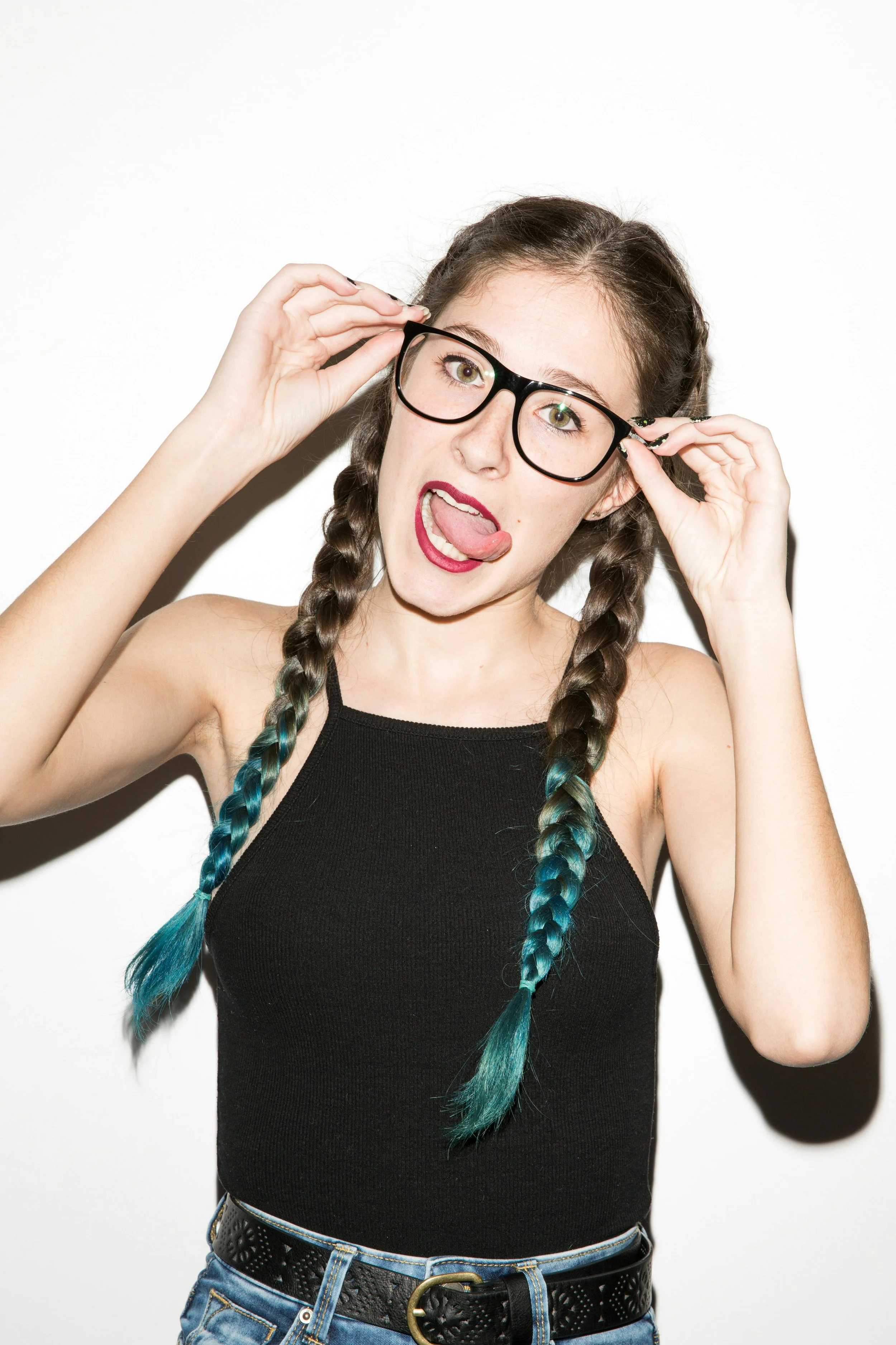 A young woman with braided hair dyed blue at the ends, wearing glasses, a black sleeveless top, and blue jeans, sticking out her tongue and making a playful face.