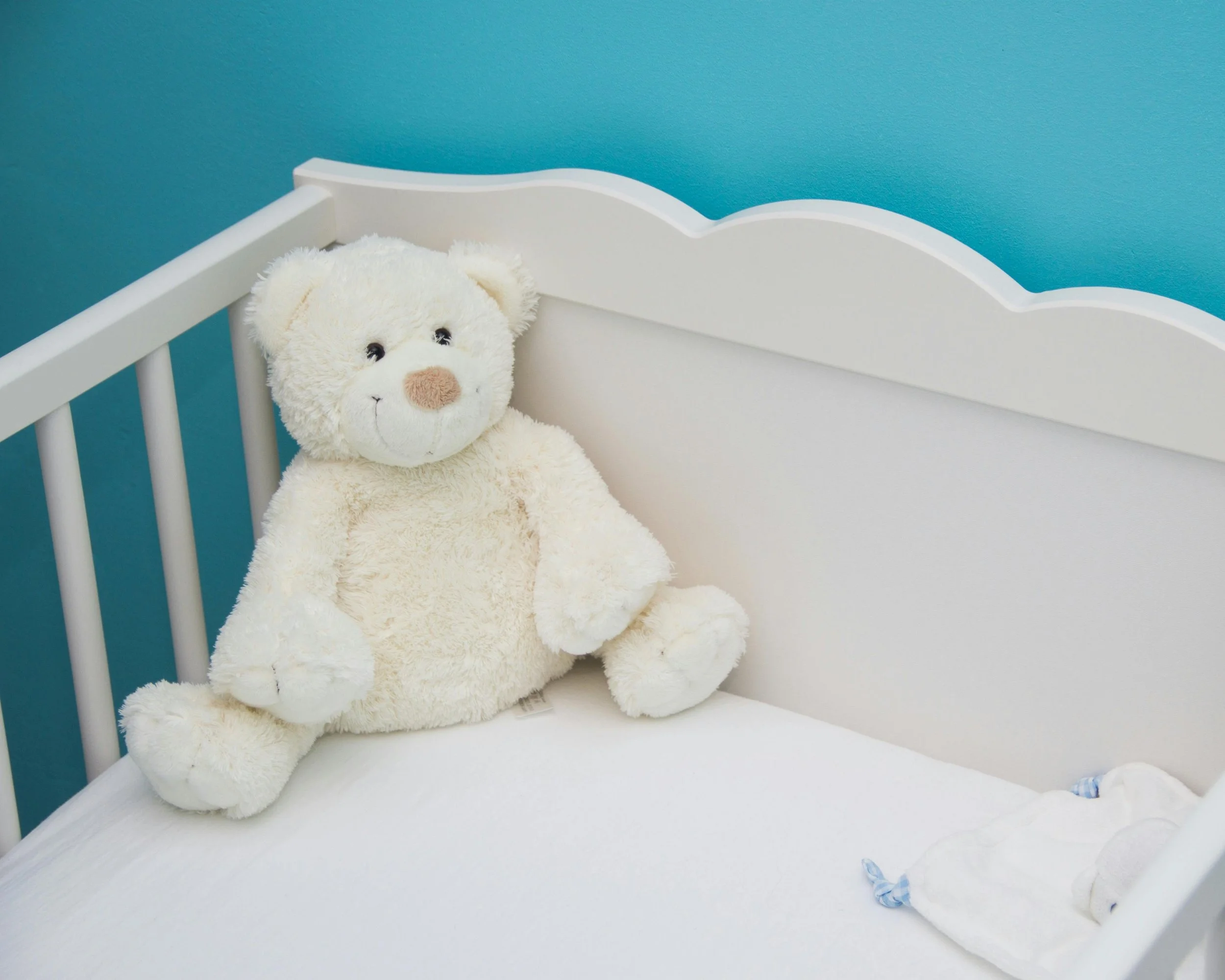 A white teddy bear with a tan nose sits on the corner of a white crib with vertical spindles, against a teal wall.