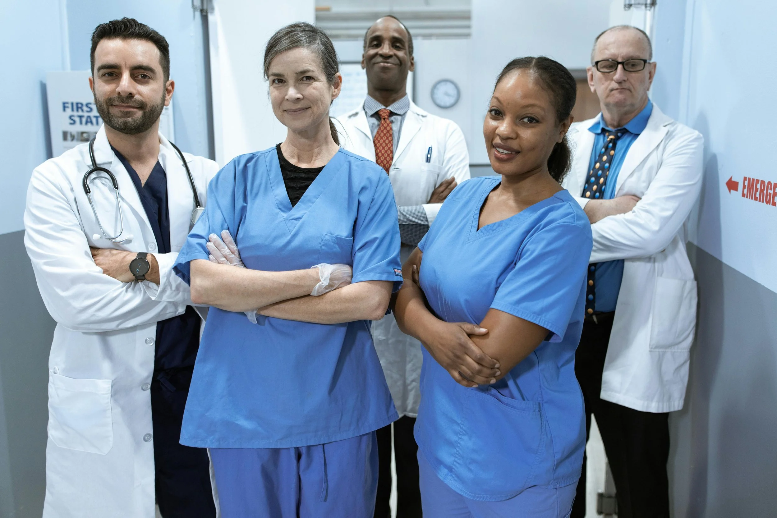 A diverse group of five medical professionals, including doctors and nurses, standing together in a hospital corridor with emergency signs and a clock in the background.