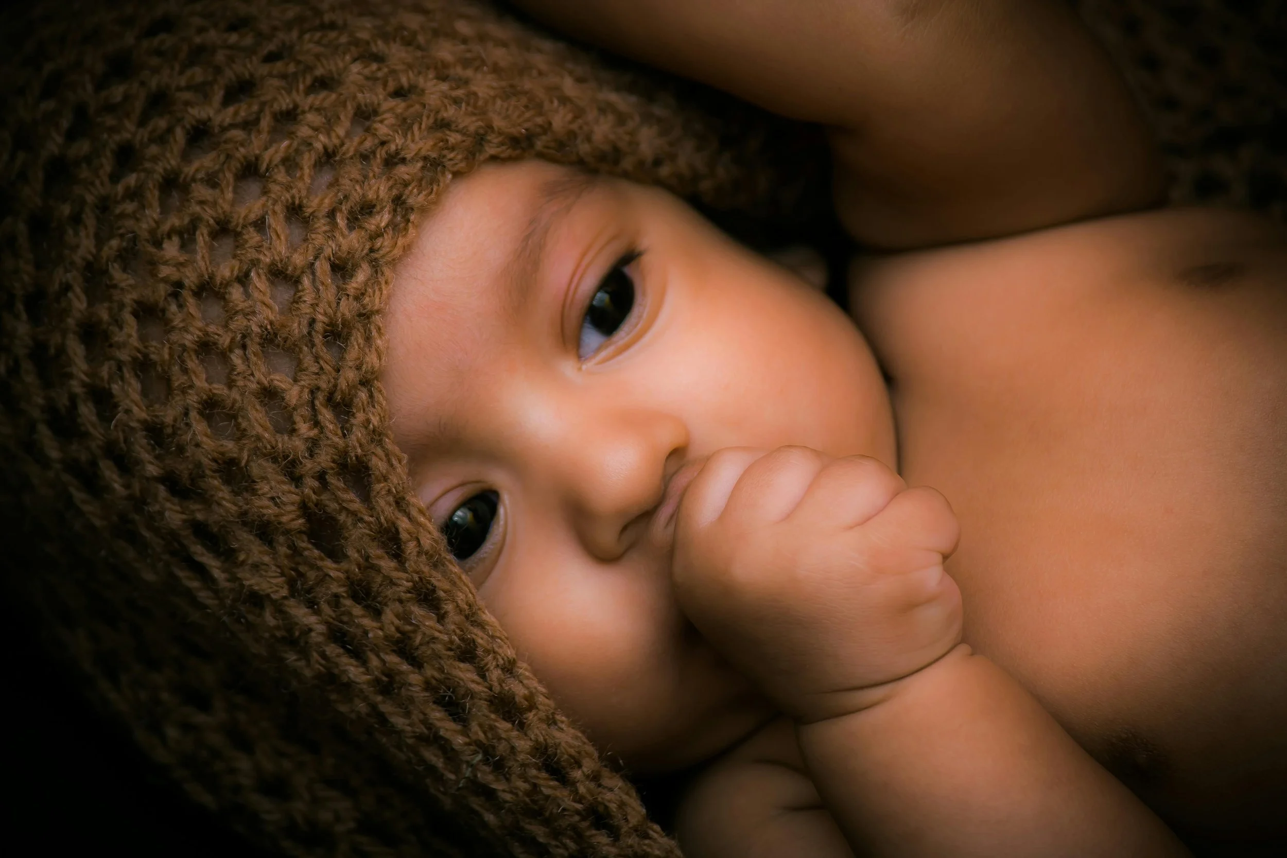 Close-up of a young child with light brown skin and dark eyes, wearing a brown knit hat, lying on their side with their hand near their face.