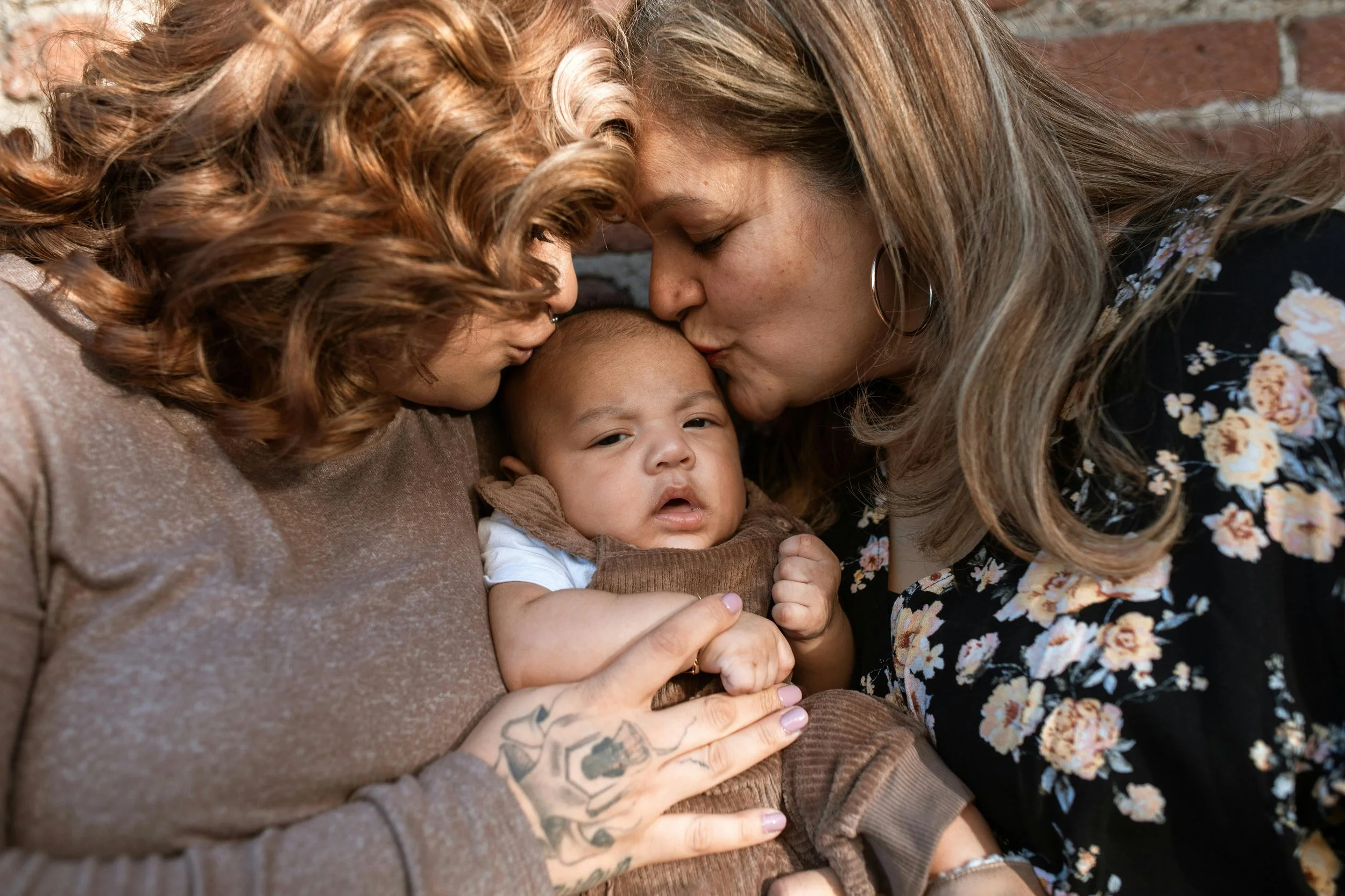 A woman with long wavy hair and hoop earrings kisses a young child on the forehead while another woman with curly hair and tattoos on her arm holds the child.
