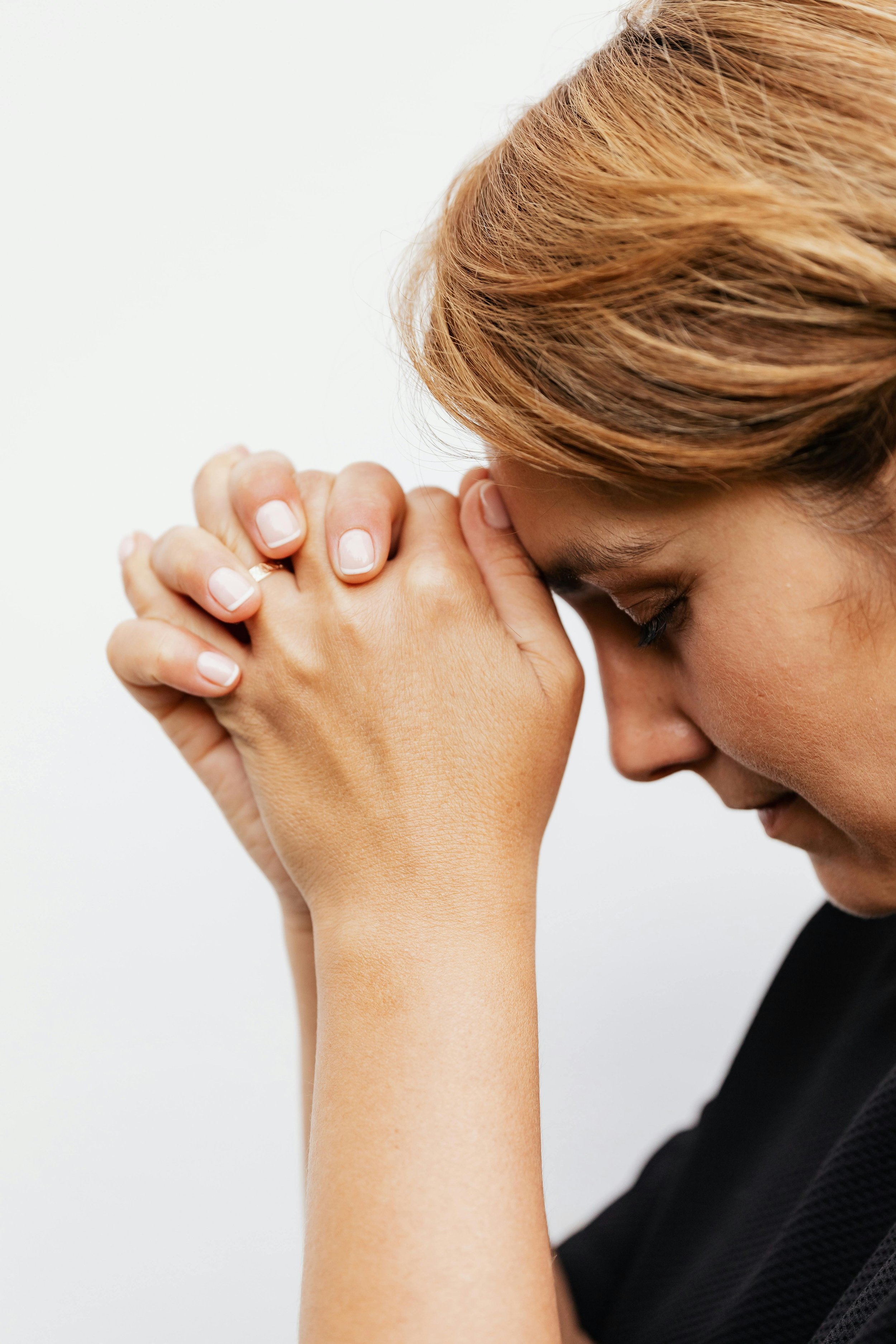 A woman with reddish hair has her eyes closed, her head bowed, and her hands clasped in front of her forehead in a prayerful or contemplative pose.