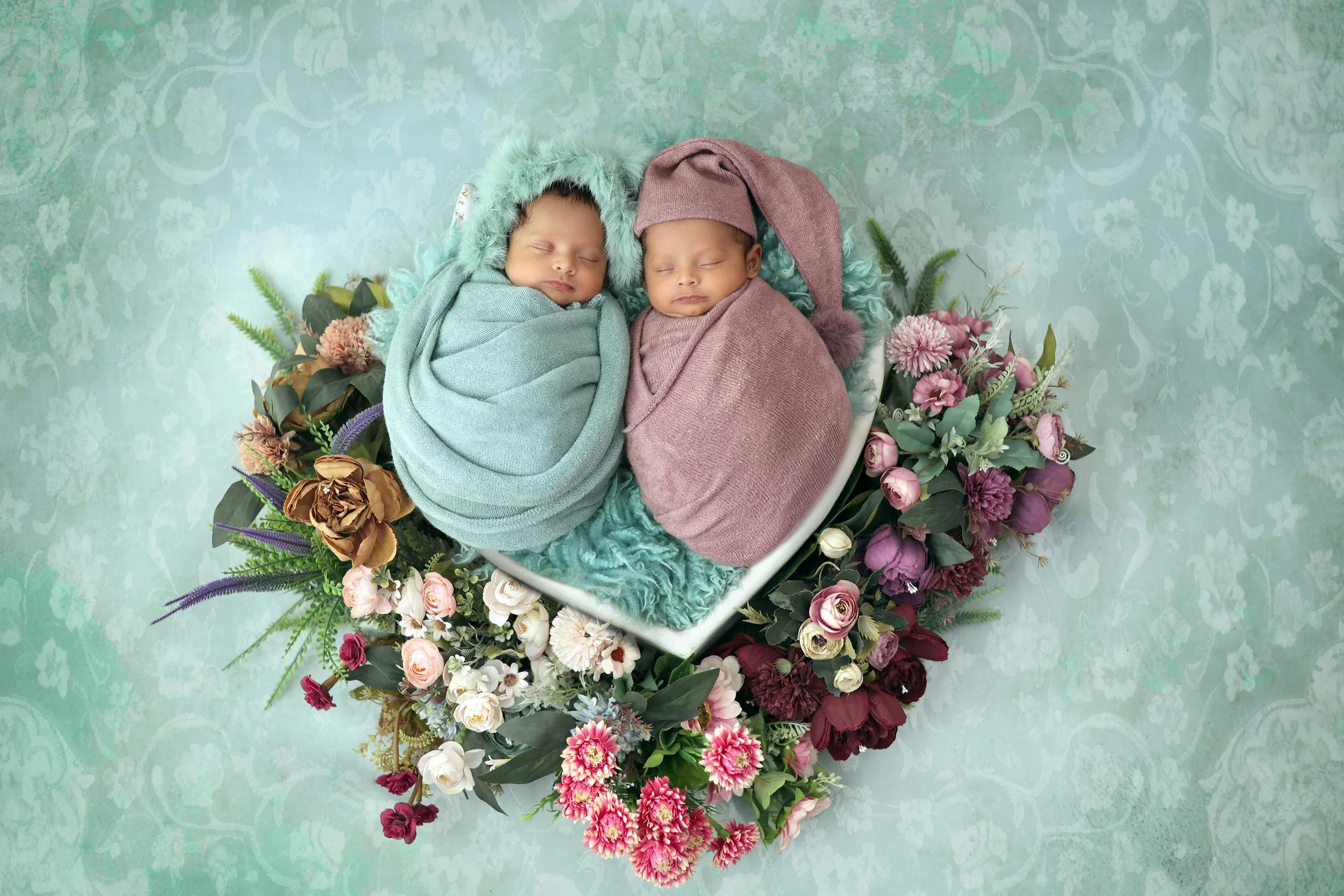 Two newborn babies wrapped in pastel-colored blankets, resting with eyes closed, lying on a bed of greenery and surrounded by a variety of pink, white, and burgundy flowers.