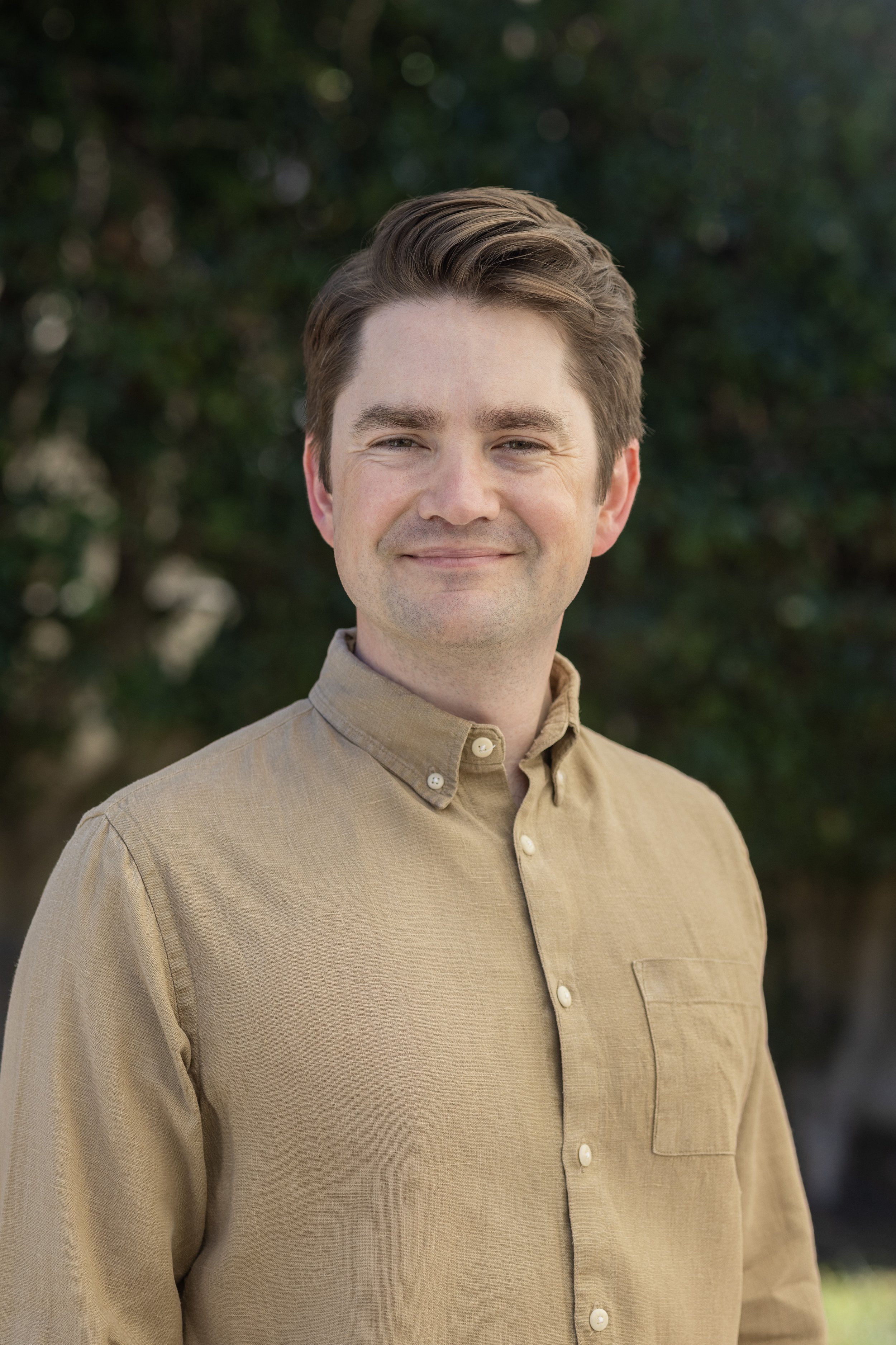 A young man with light skin, brown hair, and a slight smile, wearing a beige button-up shirt, standing outdoors with a background of blurred green foliage.