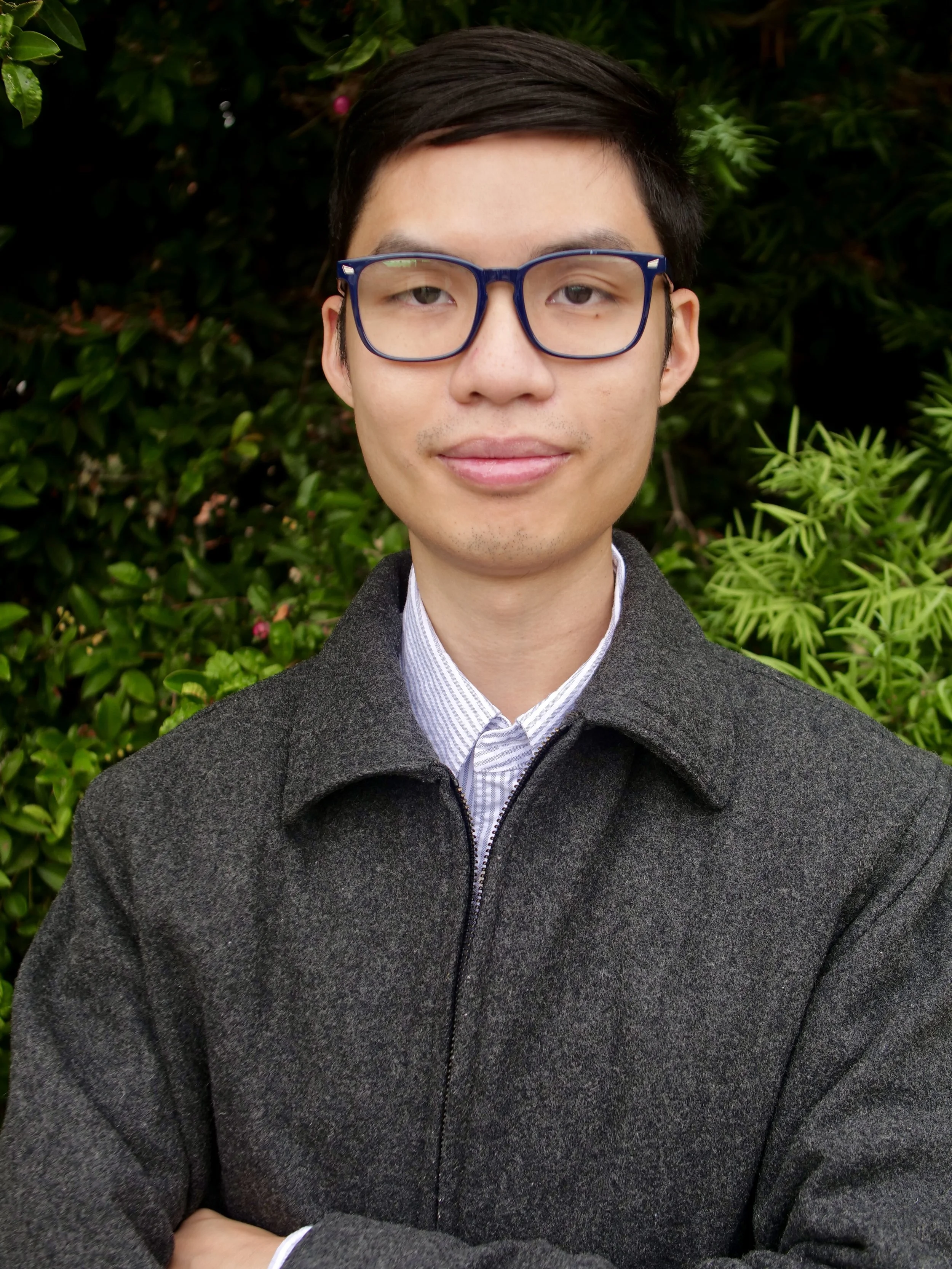 Portrait of a young man with dark hair, wearing glasses, a collared shirt, and a dark gray coat, standing outdoors with green foliage in the background.