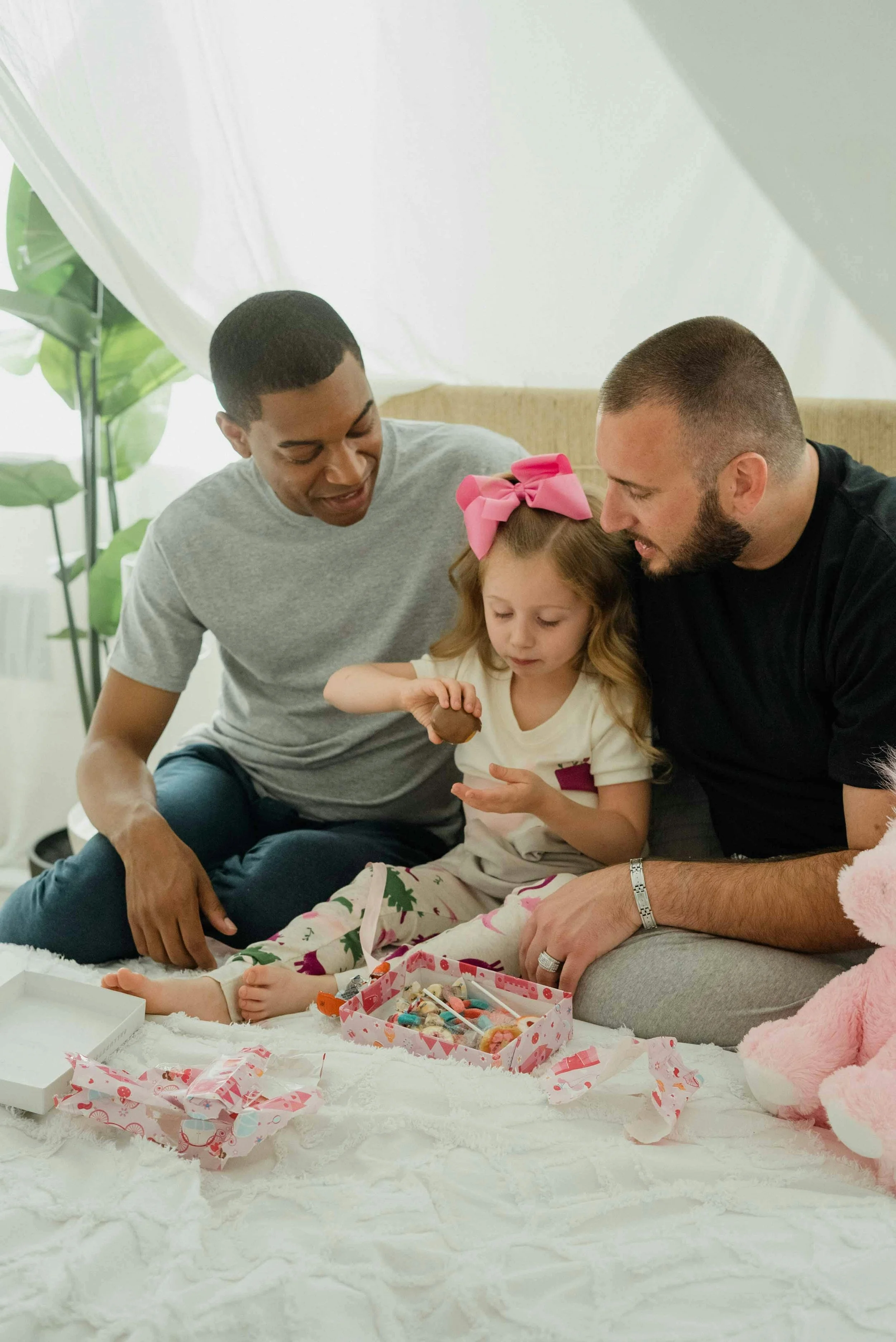 A girl with a pink bow in her hair and two men sitting on a white fluffy blanket with a box of sewing supplies in front of them, in a room with leafy plants and a white backdrop.