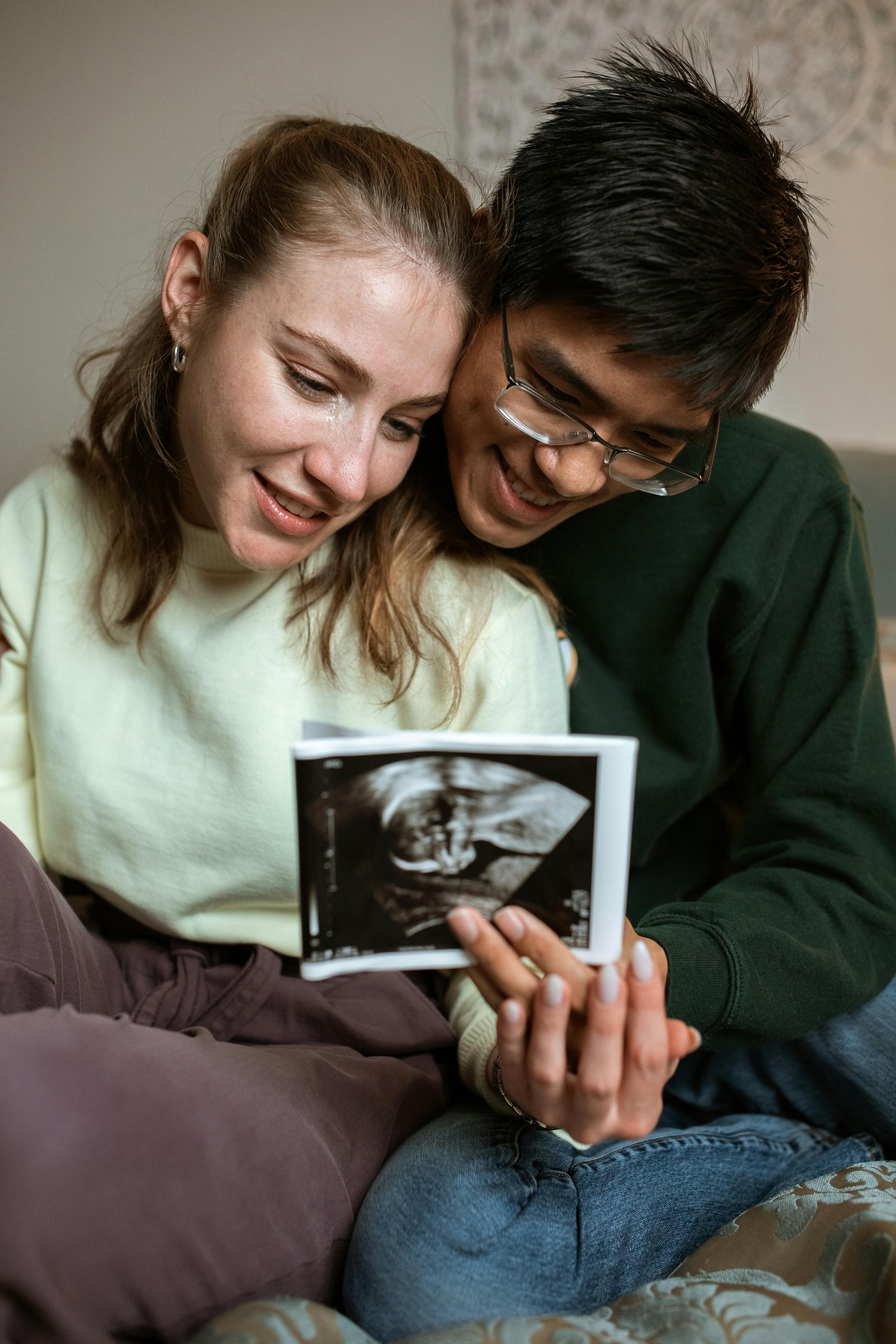 A woman and a man sitting close together, looking at an ultrasound picture of a fetus, smiling and sharing a happy moment.