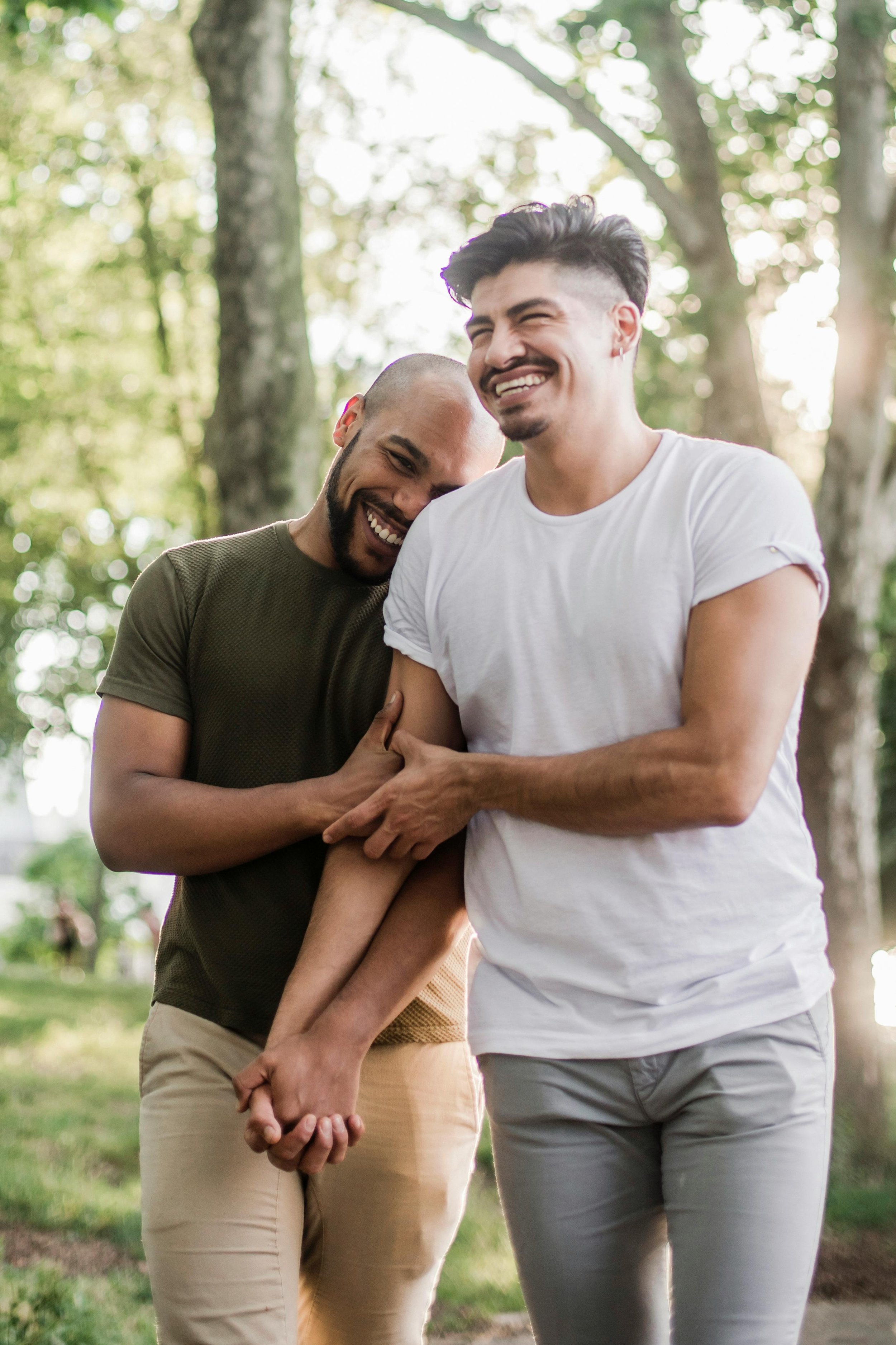 Two men are smiling and hugging in a park with trees in the background.
