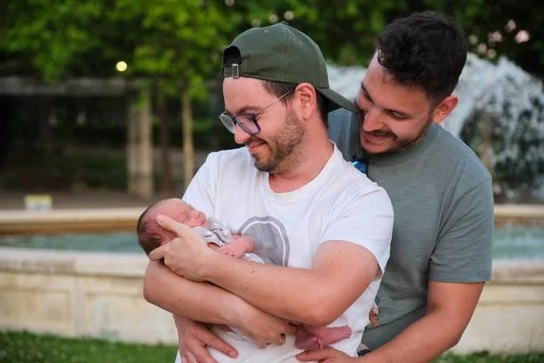 Two men smiling and holding a newborn baby outdoors in a park.
