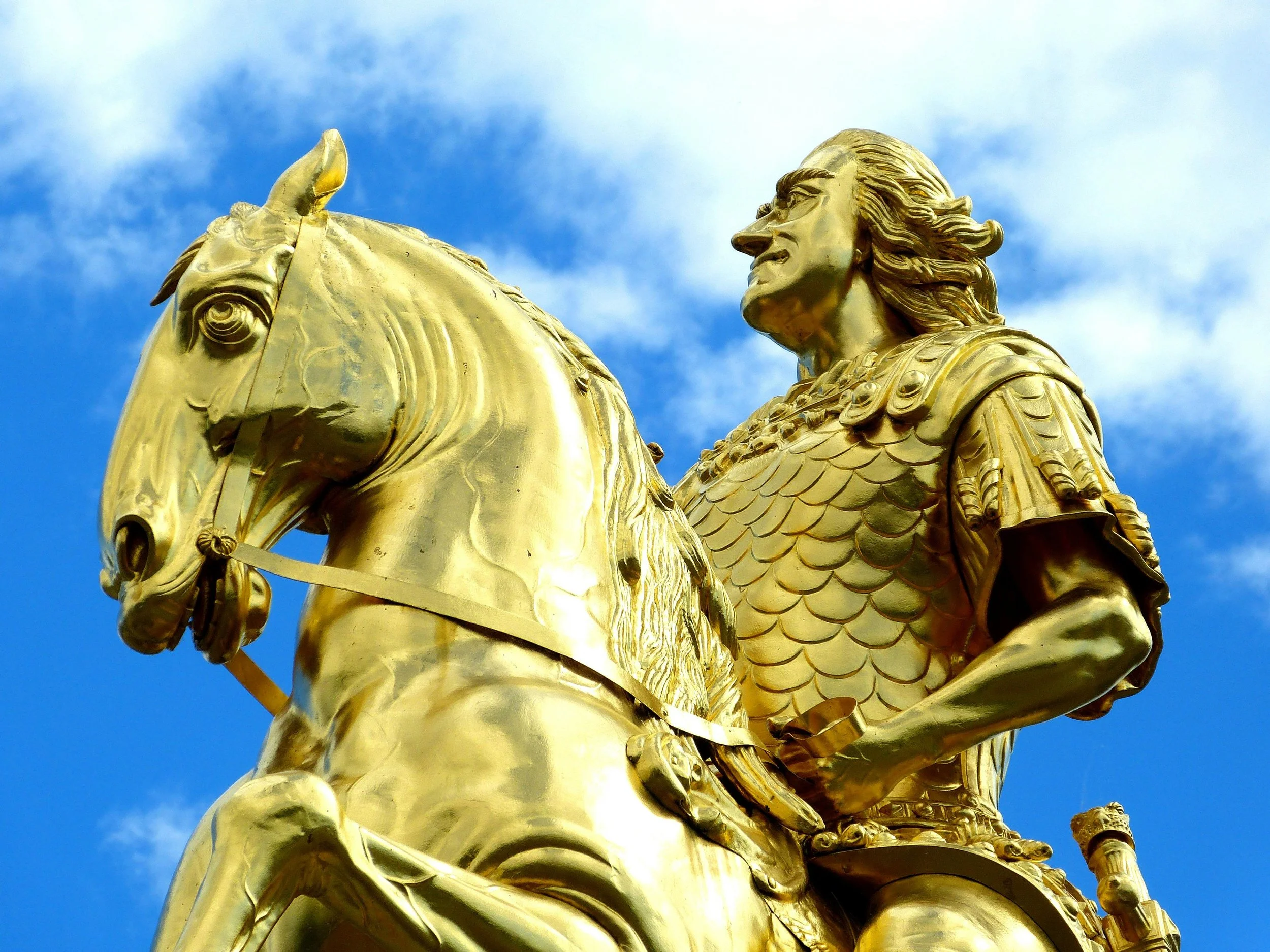 Golden statue of a man on horseback, wearing armor, with a blue sky and clouds in the background.