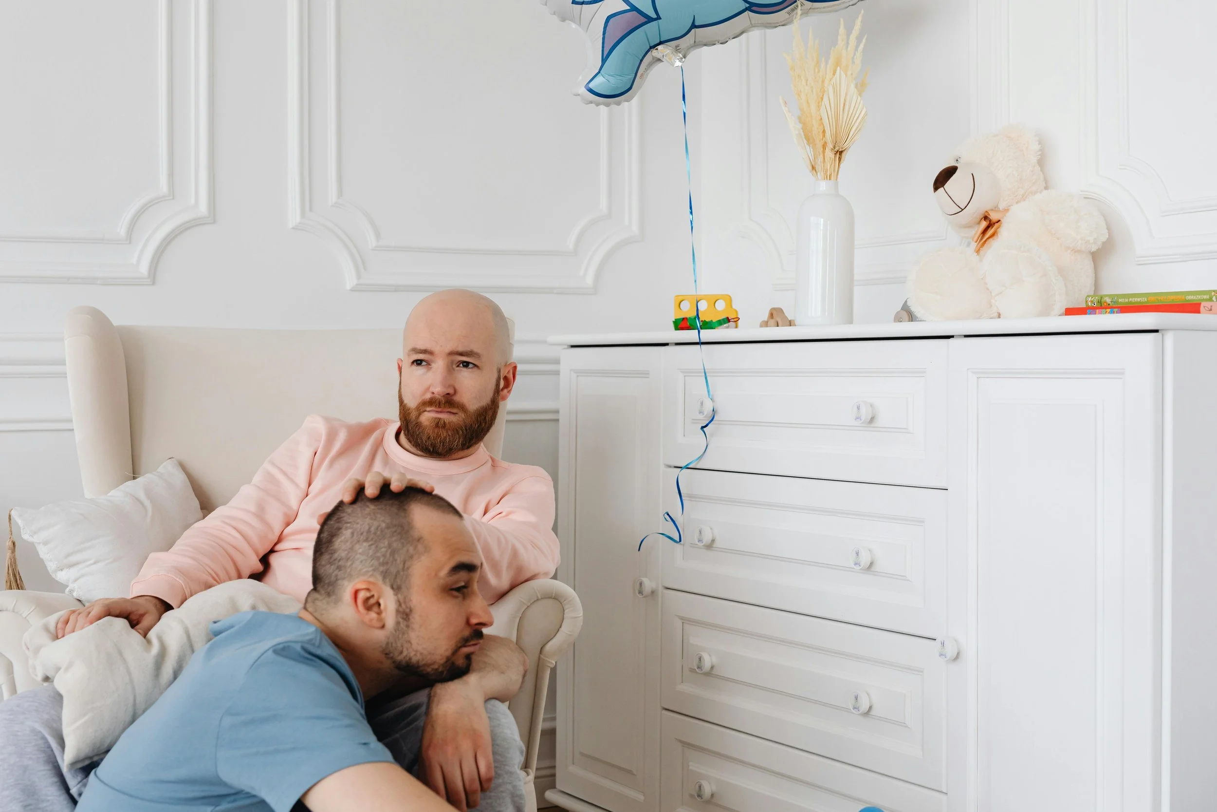 Three men sitting and lying in a white bedroom, celebrating a birthday, with a birthday balloon, teddy bear, and birthday presents in the background.