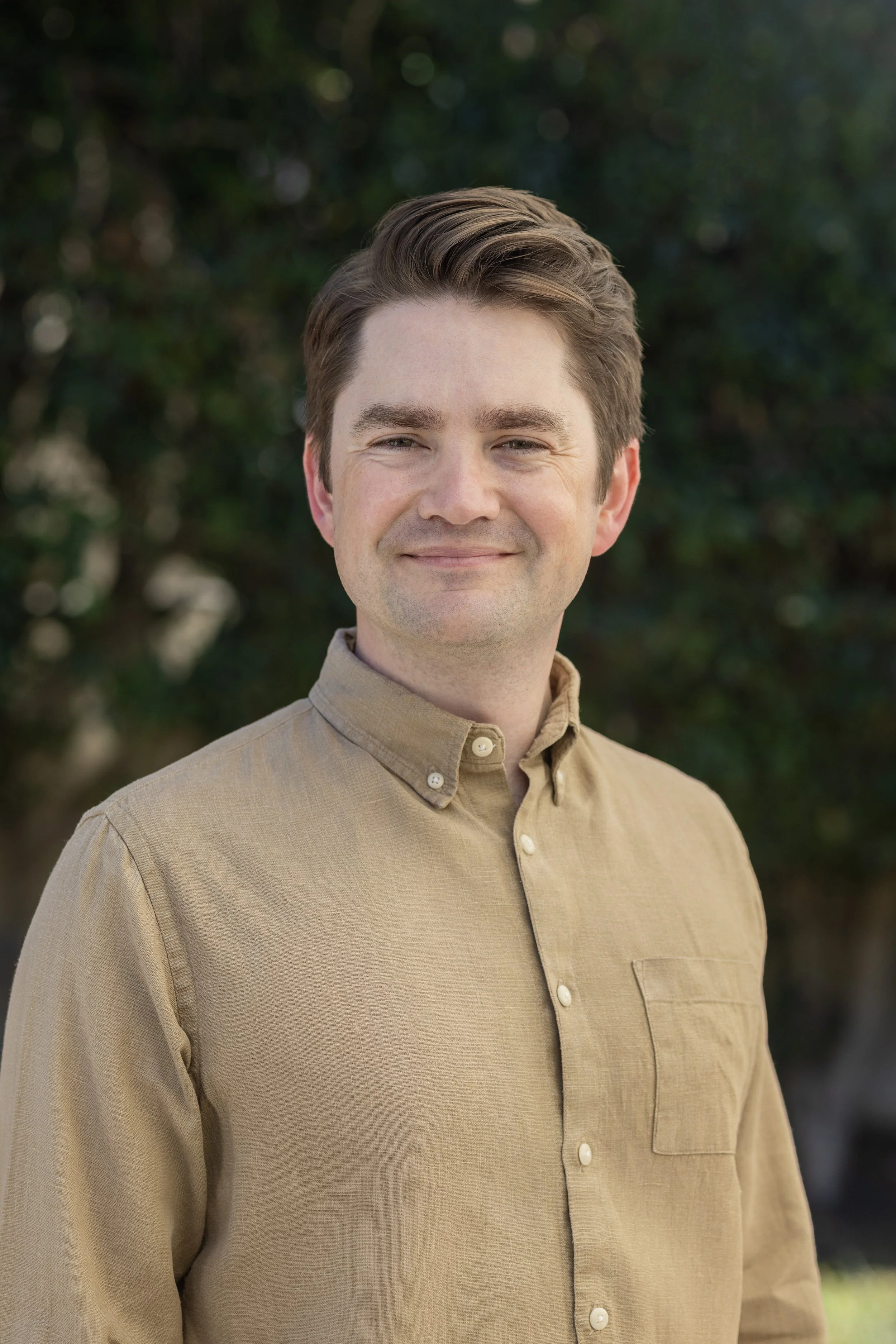 Eric Windell in a tan shirt smiling in front of green foliage.