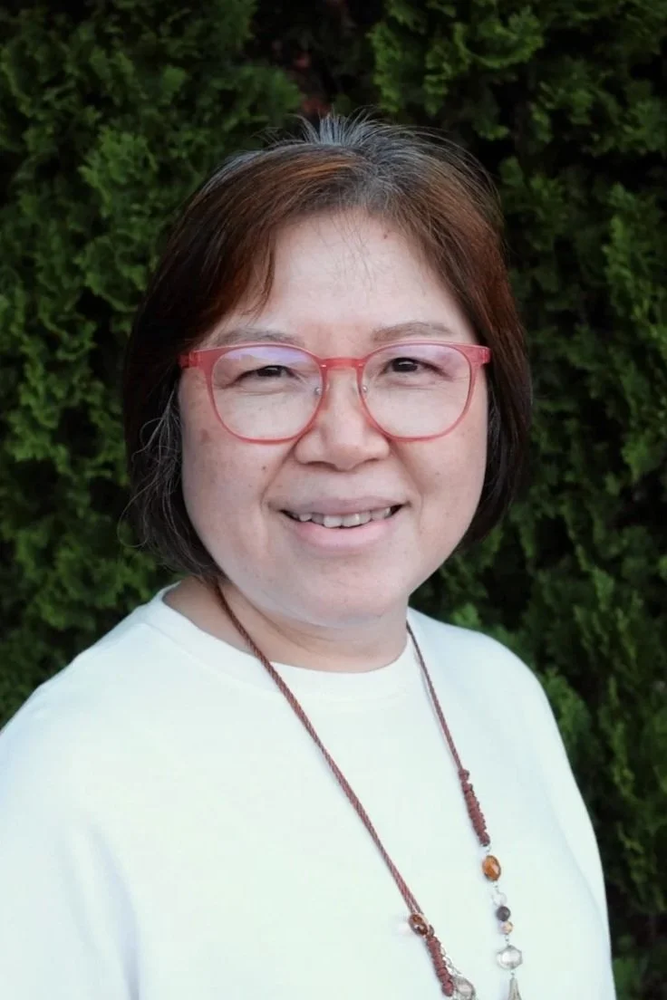 A woman with short brown hair, wearing red glasses, smiling, and dressed in a white top with a beaded necklace, standing outdoors in front of green foliage.