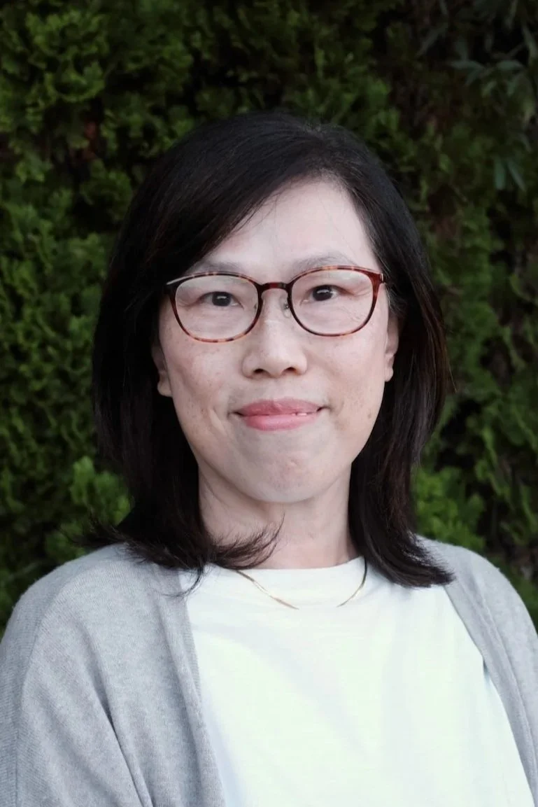 A woman with shoulder-length dark hair, glasses, and a light-colored cardigan standing outdoors in front of green foliage.