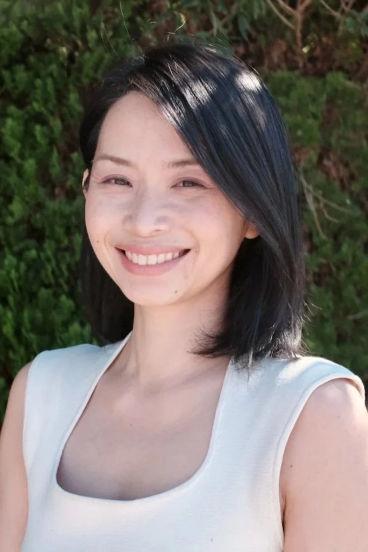 A woman with short black hair smiling outdoors in front of trees, wearing a green sleeveless top.