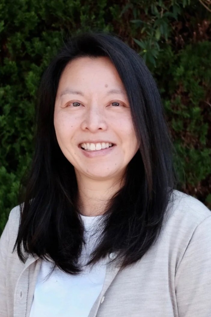 Portrait of a smiling woman with long black hair, wearing a white and navy striped shirt, a gray blazer, and a necklace, standing outdoors with green foliage in the background.