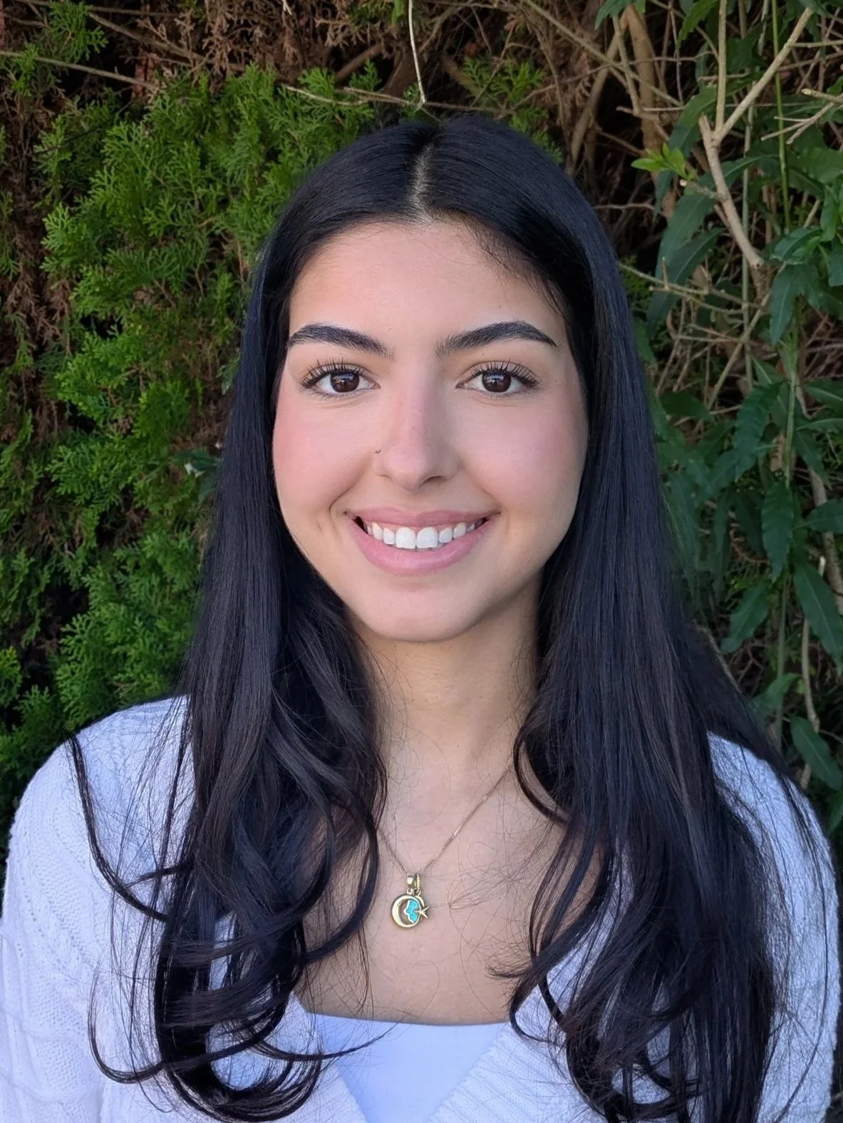 A young woman with long dark hair and earrings, smiling in front of a leafy green hedge.