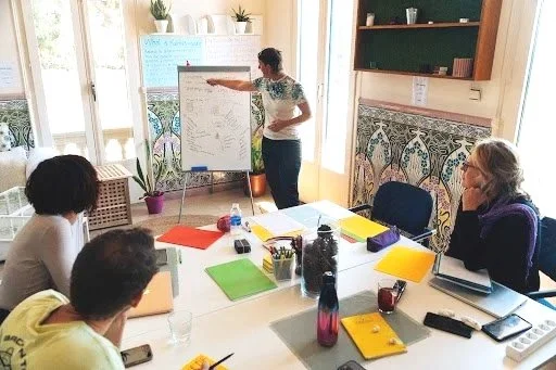 A group of four people attending a meeting or workshop in a brightly lit room with large windows. One person is standing and presenting on a flip chart with notes and diagrams. The others are seated at a table with notebooks, water bottles, and electronic devices, listening attentively.