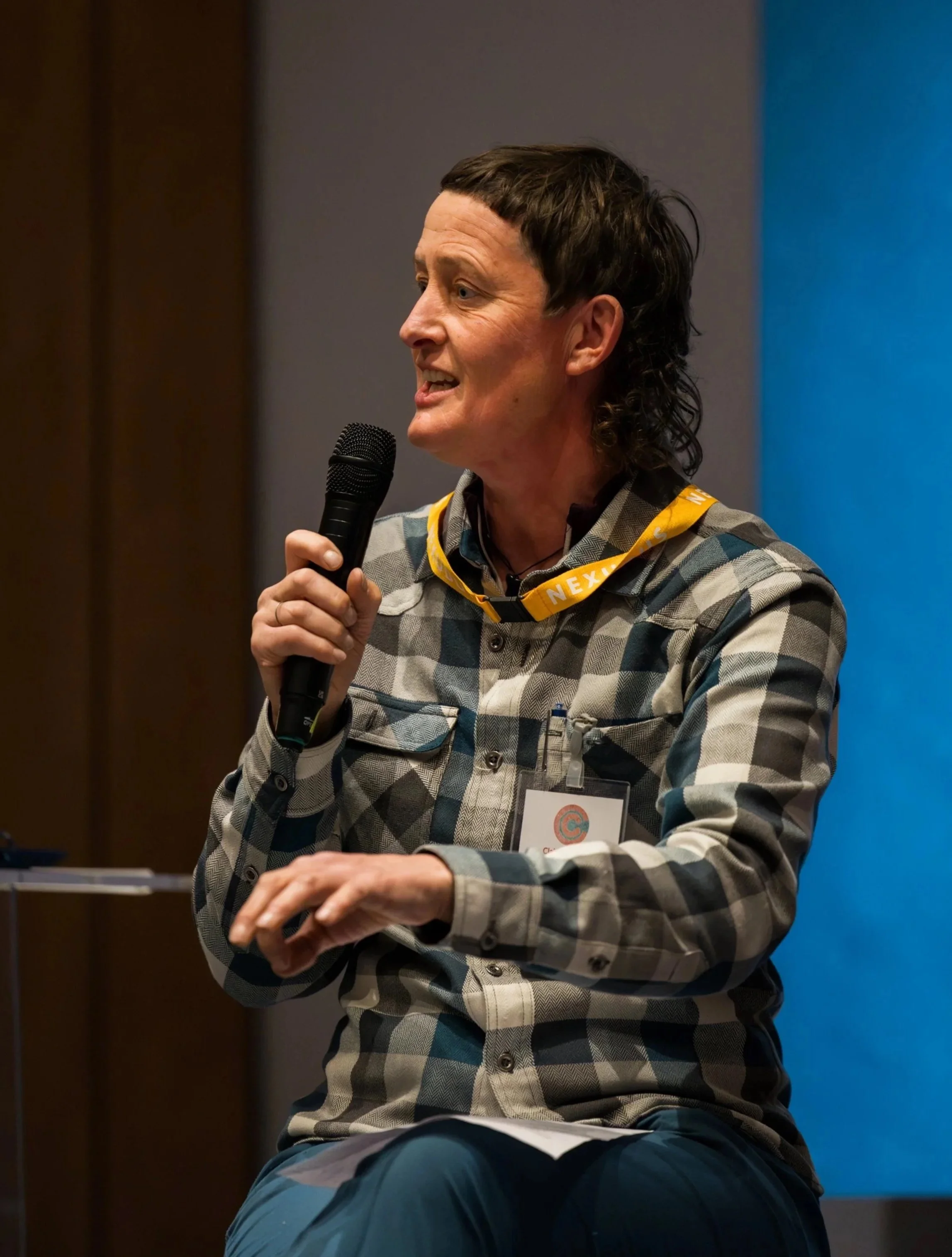 A woman with short dark hair and a checkered shirt speaking into a microphone during a presentation at an indoor event.