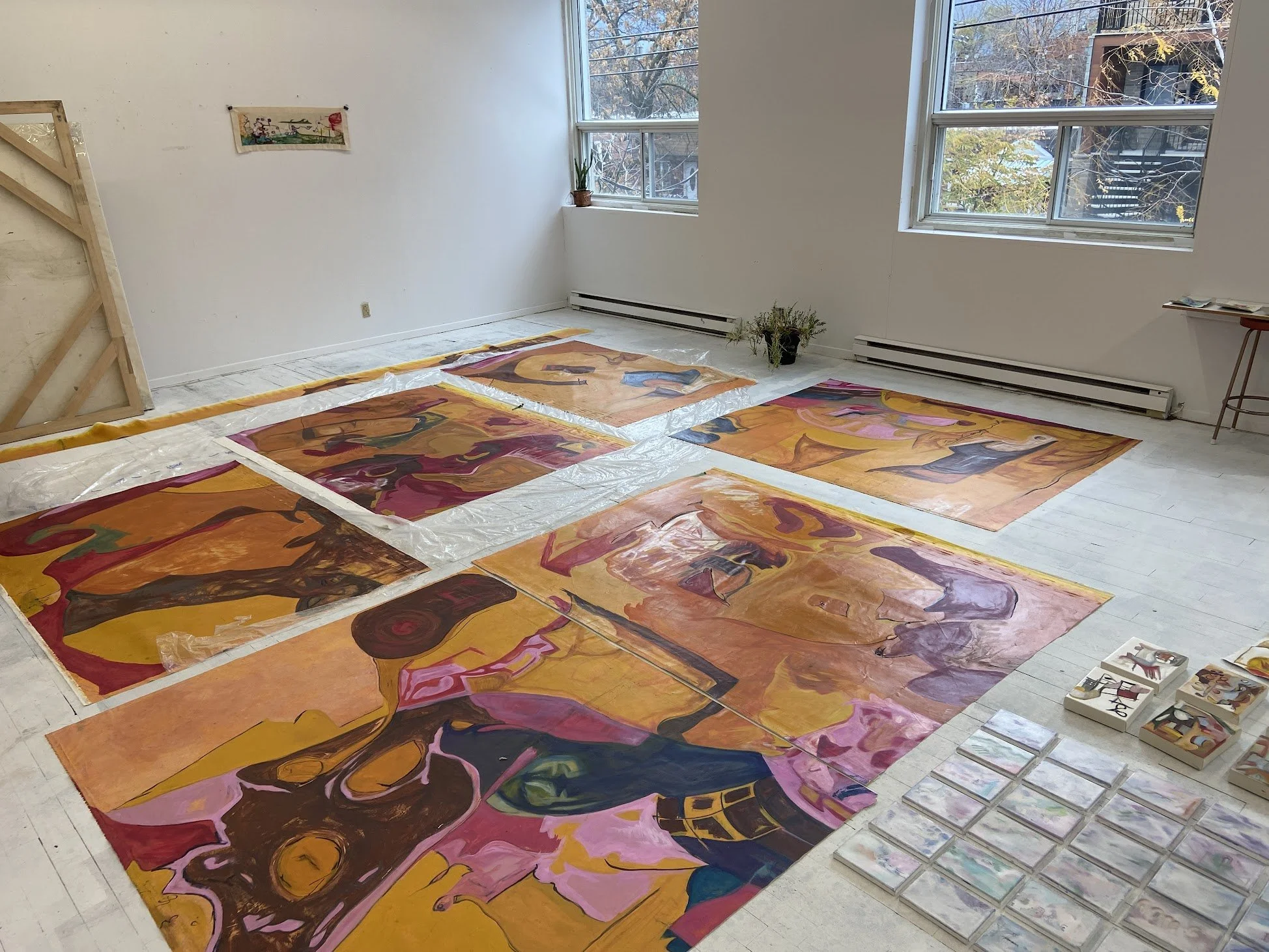 An art studio with large abstract paintings laid out on the white wooden floor, some with plastic sheets underneath. There are windows letting in natural light, a small plant on the windowsill, and a few art supplies on the side.