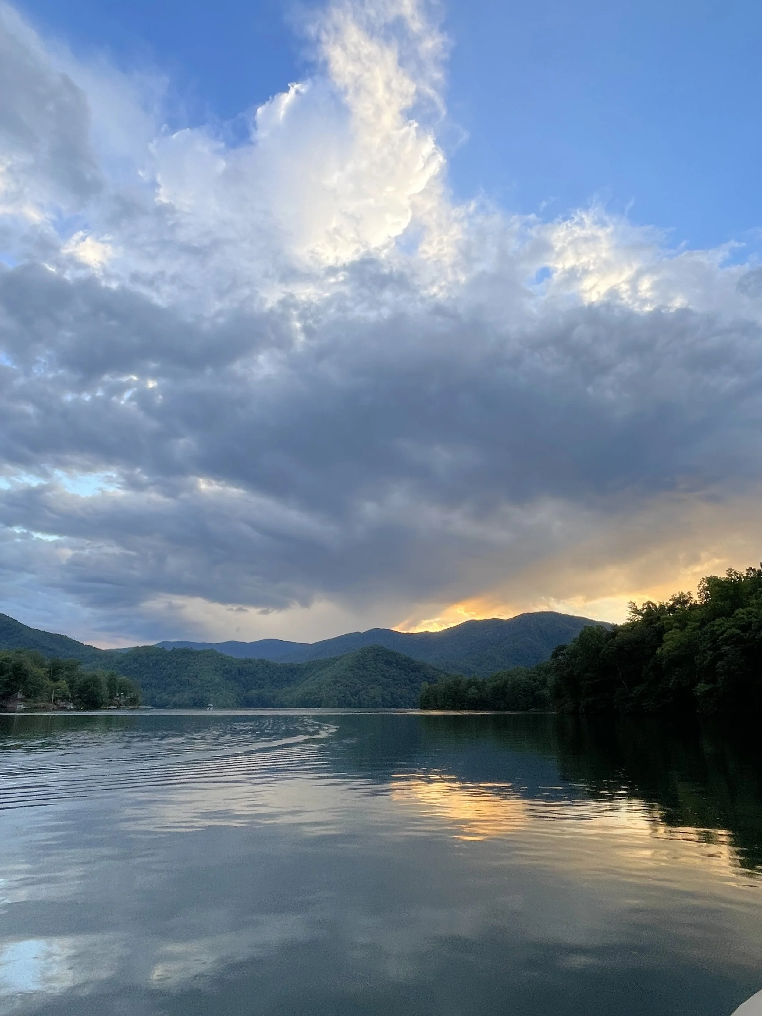 A serene lake surrounded by green mountains under a partly cloudy sky at sunset.