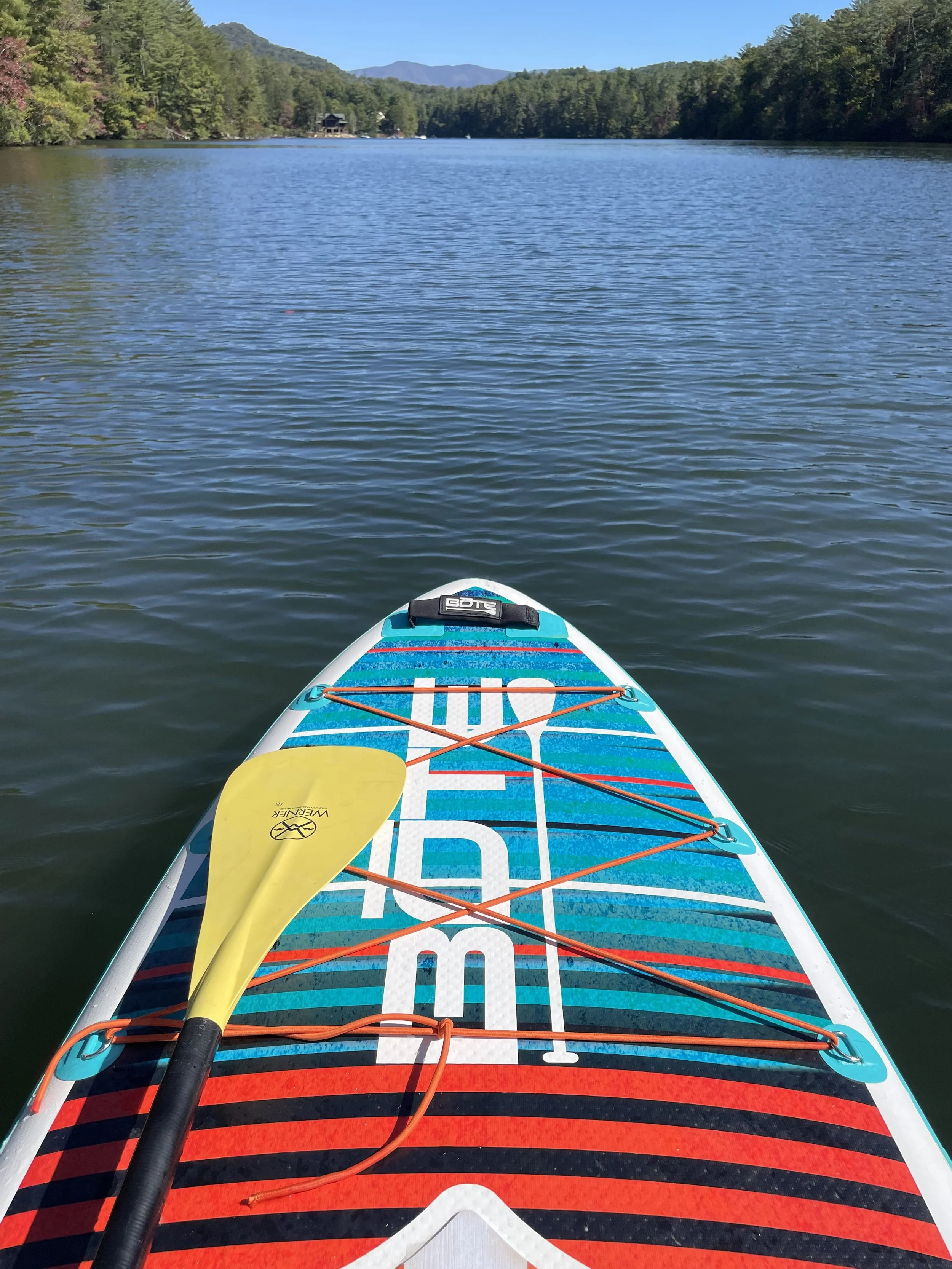 View from a paddleboard on a calm lake, with a yellow paddle lying across the board. The lake is surrounded by green forested hills and distant mountains under a clear blue sky.