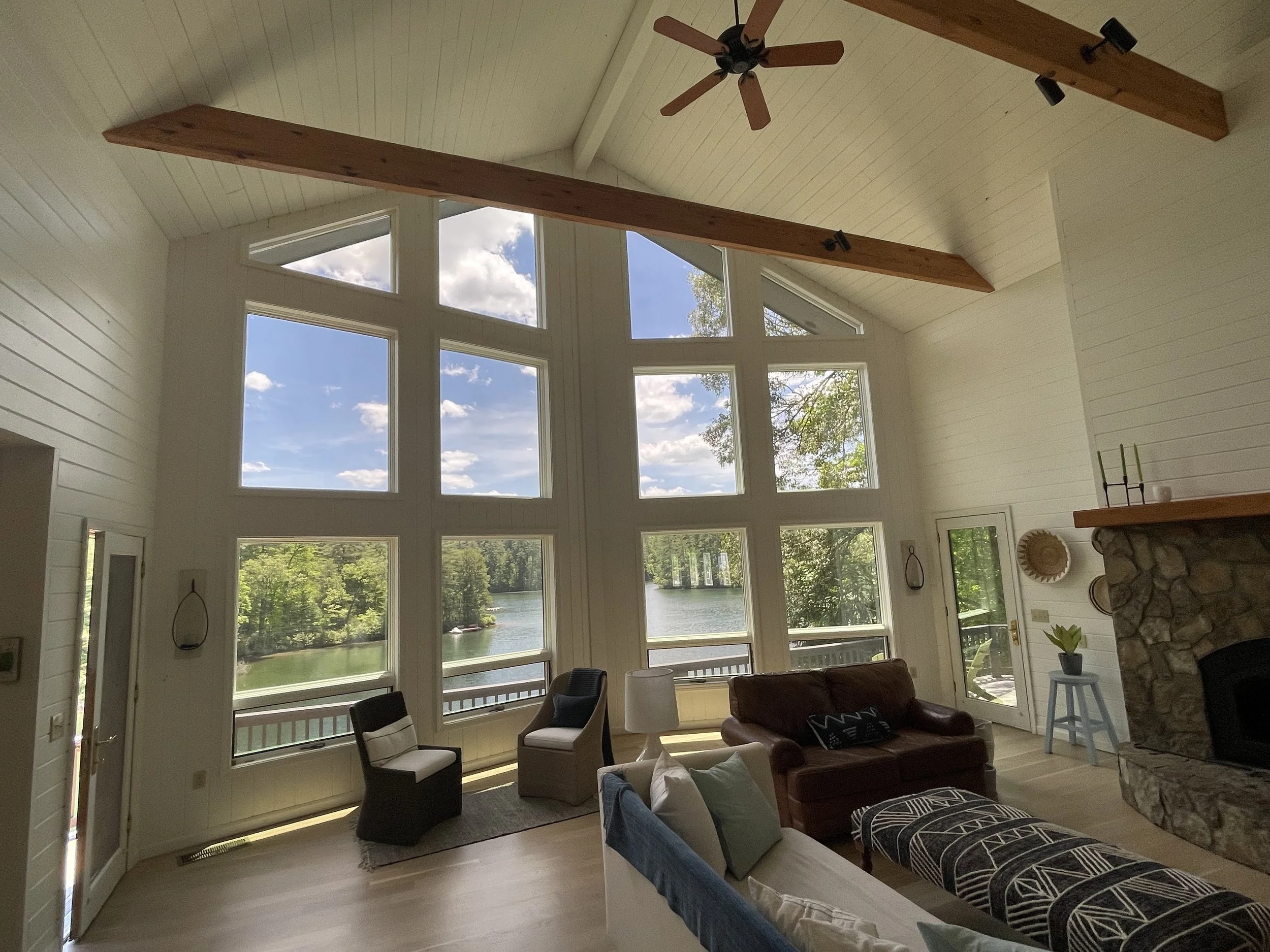 Living room with large window and lake view, high vaulted ceiling with wooden beams, ceiling fan, fireplace, and furniture including a leather sofa, armchairs, and decorative items.