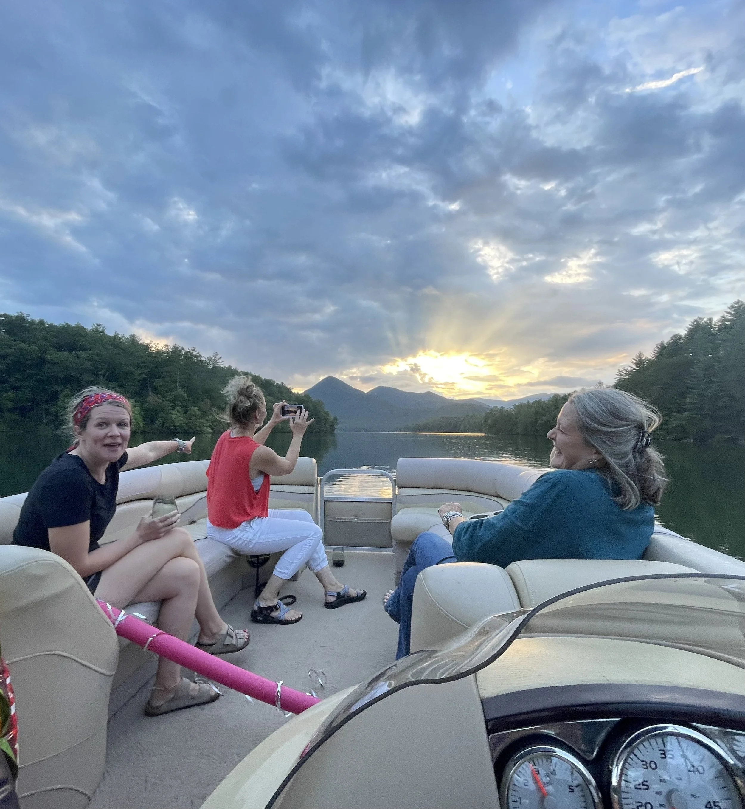 Three women on a boat at sunset, with mountains and a lake in the background. One woman is taking a photo, another is sitting and talking, and the third is smiling and holding a drink.