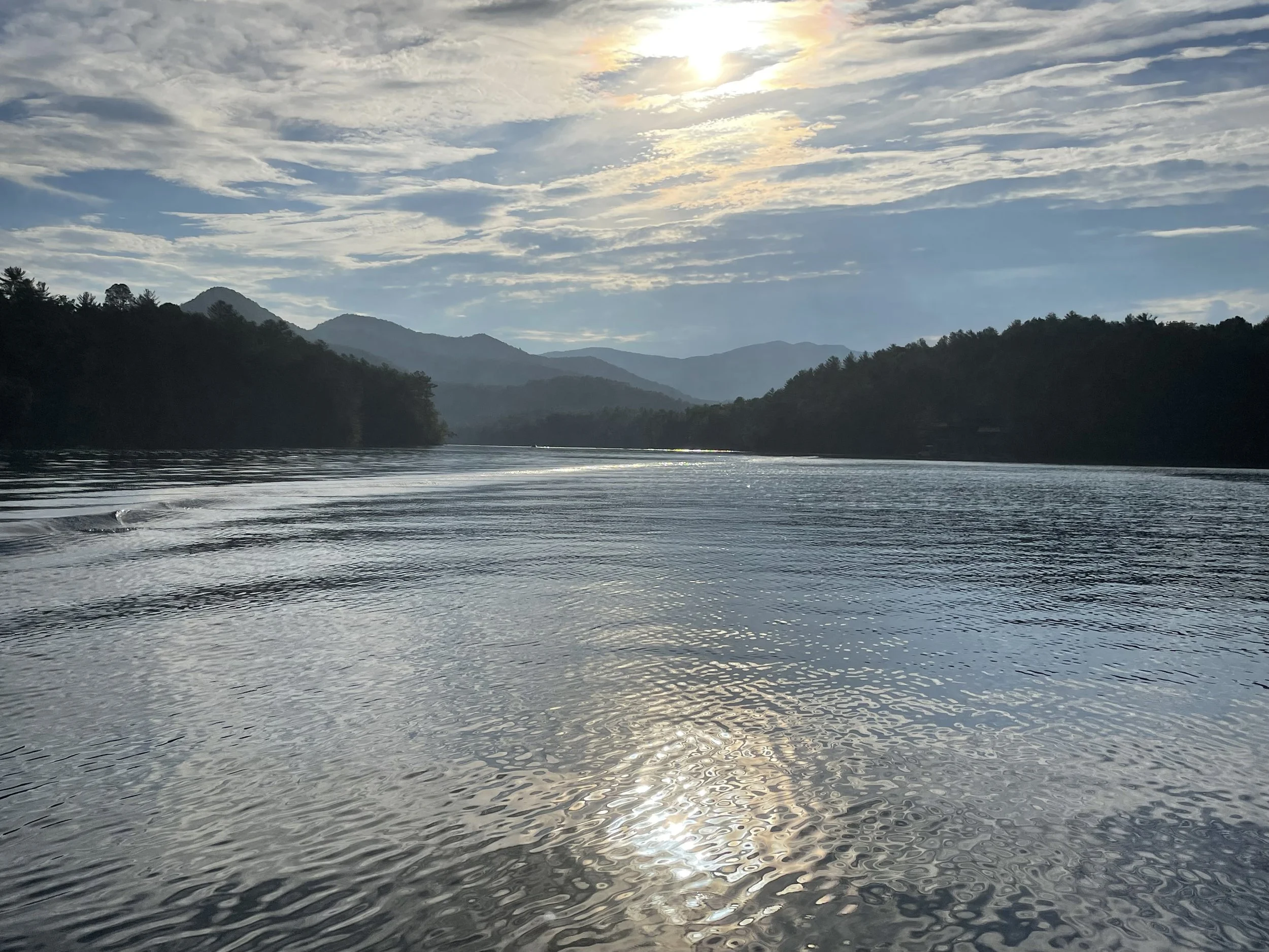 Scenic view of a lake with rippling water reflecting sunlight, surrounded by forested hills and distant mountains under a partly cloudy sky.