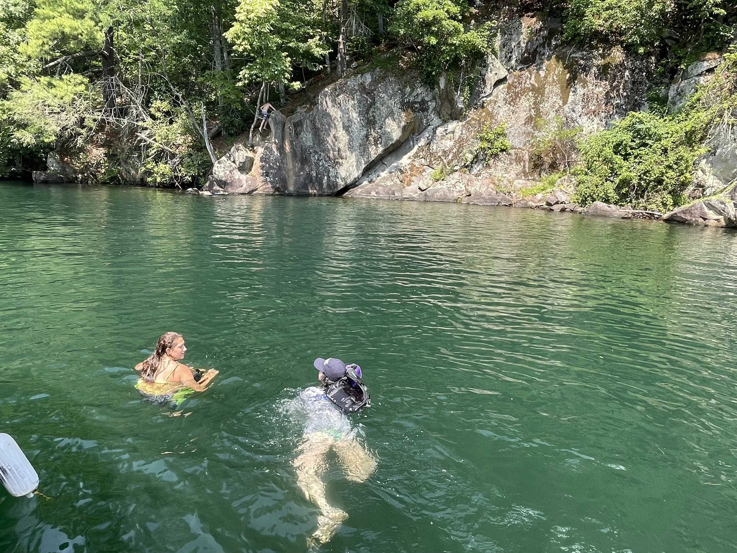 Two individuals swimming in a green river near a rocky, forested shoreline with a person climbing rocks in the background.
