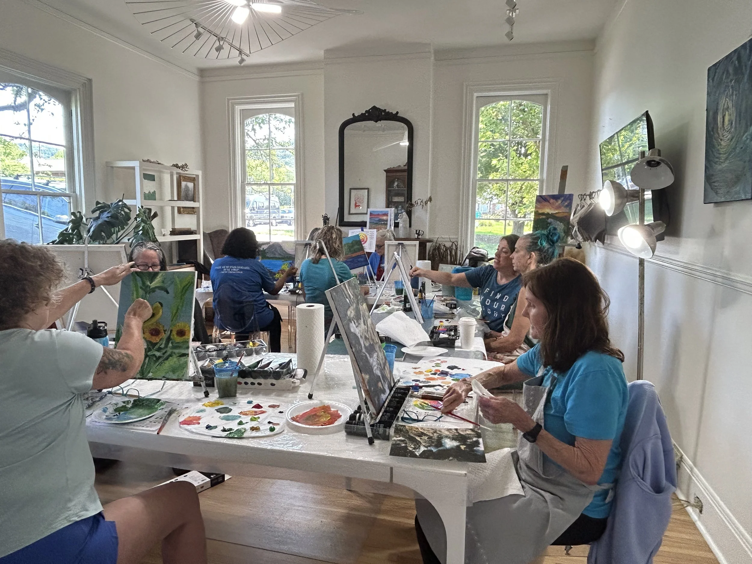 People participating in a group painting class in a bright, spacious room with large windows, white walls, and artworks on easels.