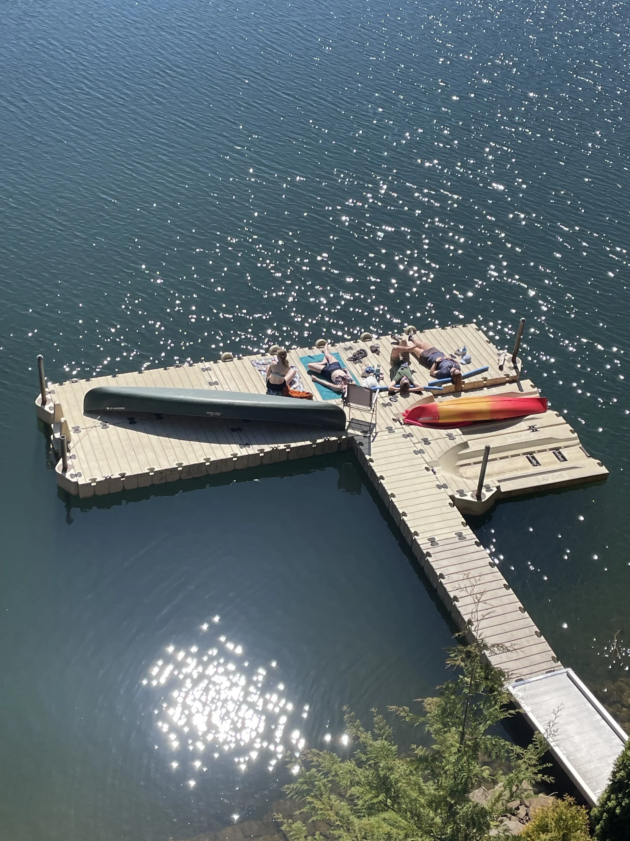 People sunbathing and relaxing on a wooden dock with kayaks and a canoe, surrounded by a calm body of water with sunlight reflecting off the surface.